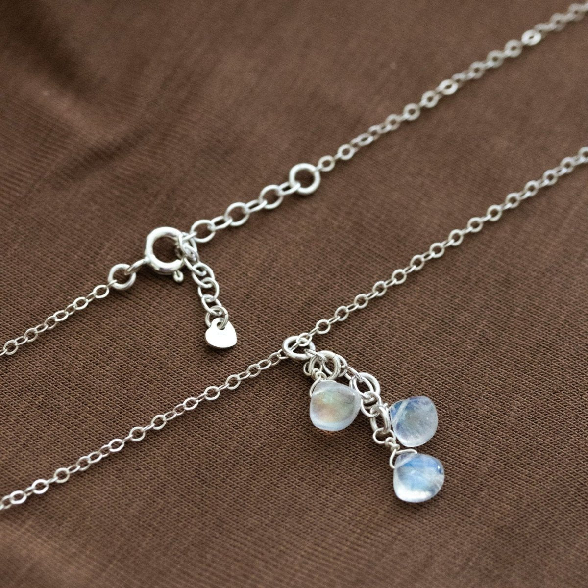 Eine dünne Silberkette mit drei untereinander angebrachten Mondstein-Tropfen liegt auf braunem Stoff.
