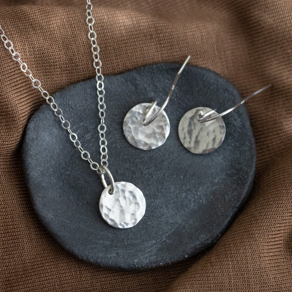 Auf einer grauen Steinschale liegt ein silbernes Schmuckset, bestehend aus einer Kette und Ohrringen mit jeweils einem runden, gehämmerten Plättchen-Anhänger.