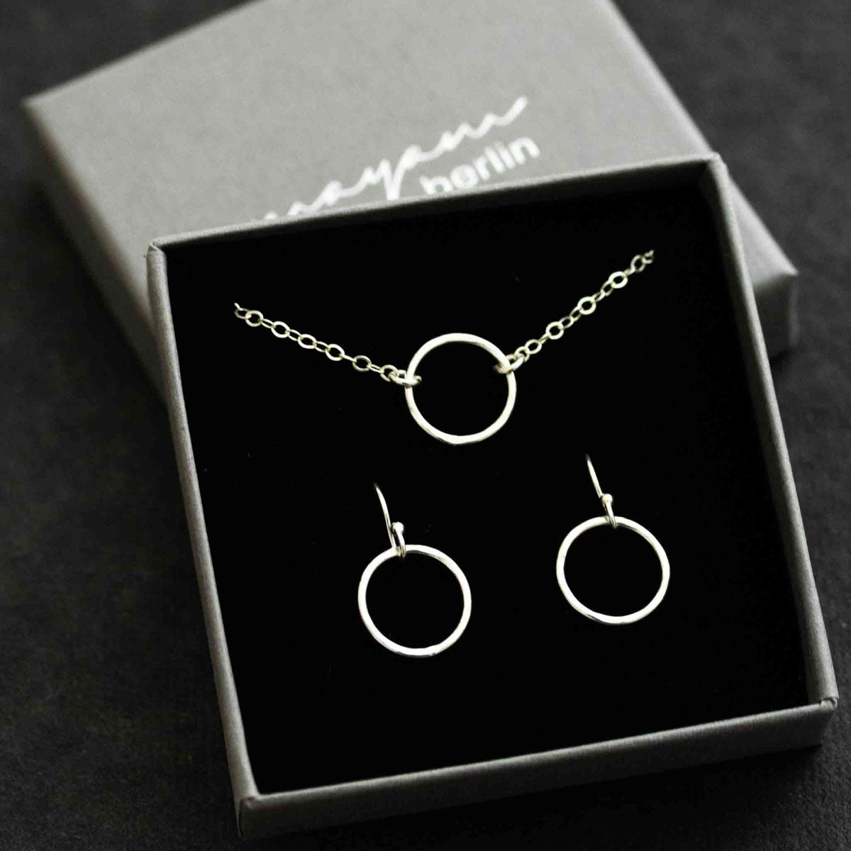 Ein Schmuckset - bestehend aus einer feinen Silberkette mit offenem Ring in der Mitte und passenden Ohrringen - ist in ein offenes Schmucketui eingehängt.