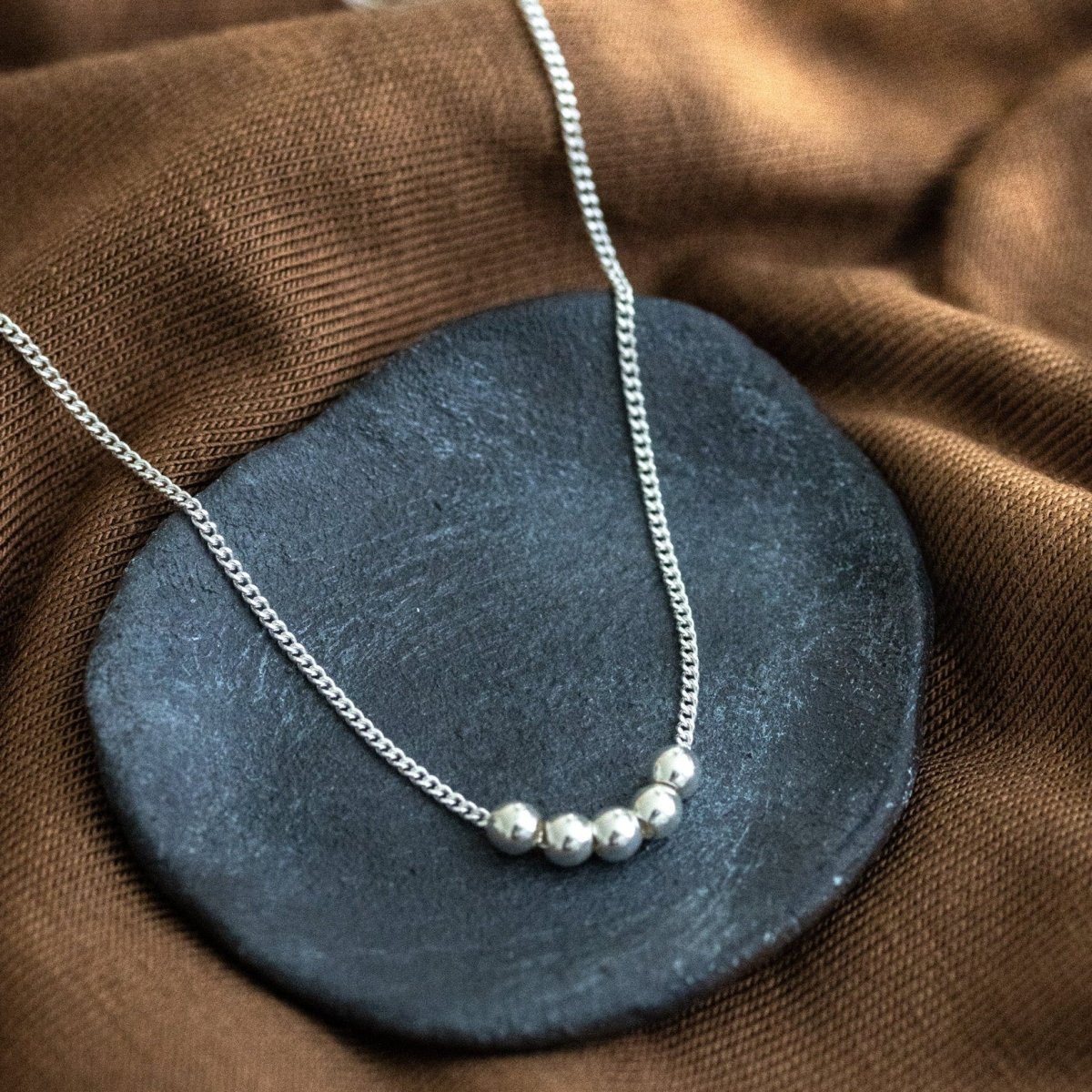 Eine silberne Panzerkette mit einer Reihe silberner Perlen in der Mitte liegt auf einem schwarzen Schälchen, das auf braunem Stoff steht.