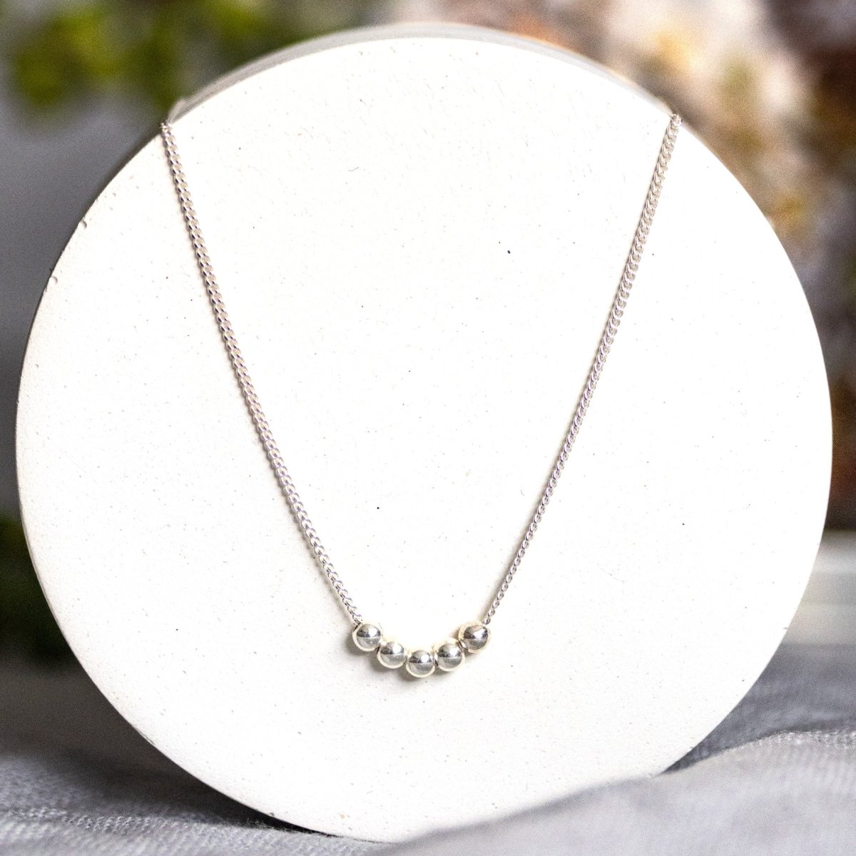 Eine silberne Halskette mit fünf aufgezogenen Silberperlen hängt an einem runden, weißen Hintergrund.