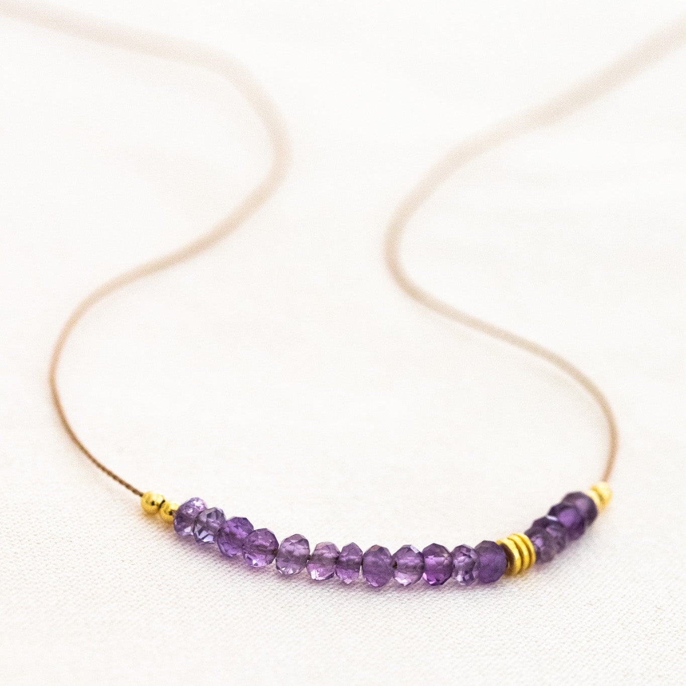 Eine dünne Seidenkette mit einem Amethyst-Cluster und goldenen Zierperlen liegt auf weißem Untergrund.