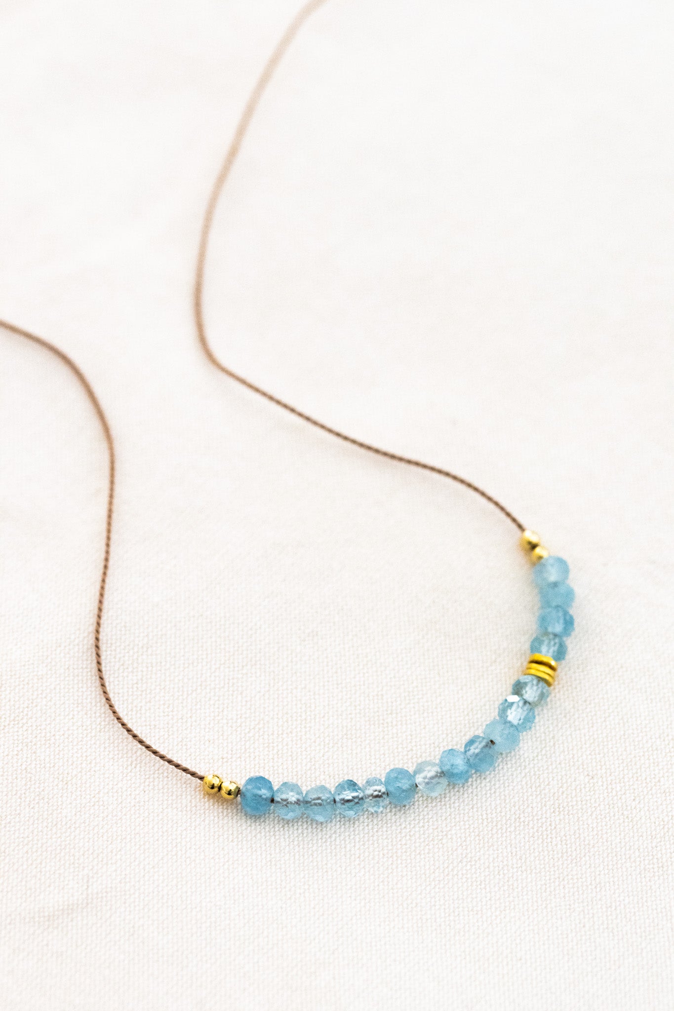 Eine Halskette aus feiner Seidenschnur mit einem Aquamarin-Cluster und goldenen Zierperlen liegt auf weißem Untergrund.
