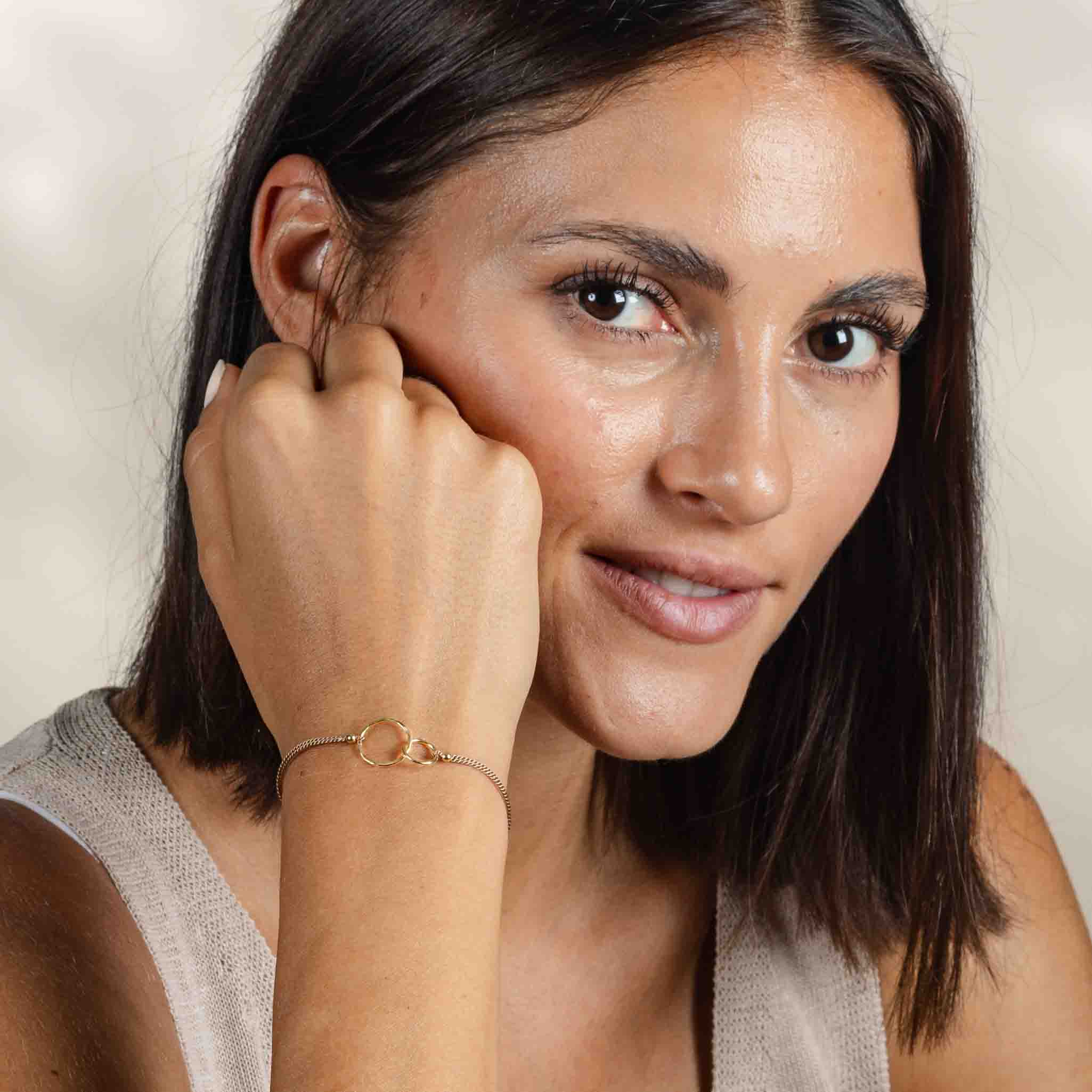 Am Handgelenk des Models hängt ein feines Seidenarmband mit zwei goldenen, ineinander verschlungenen Ringen in der Mitte.