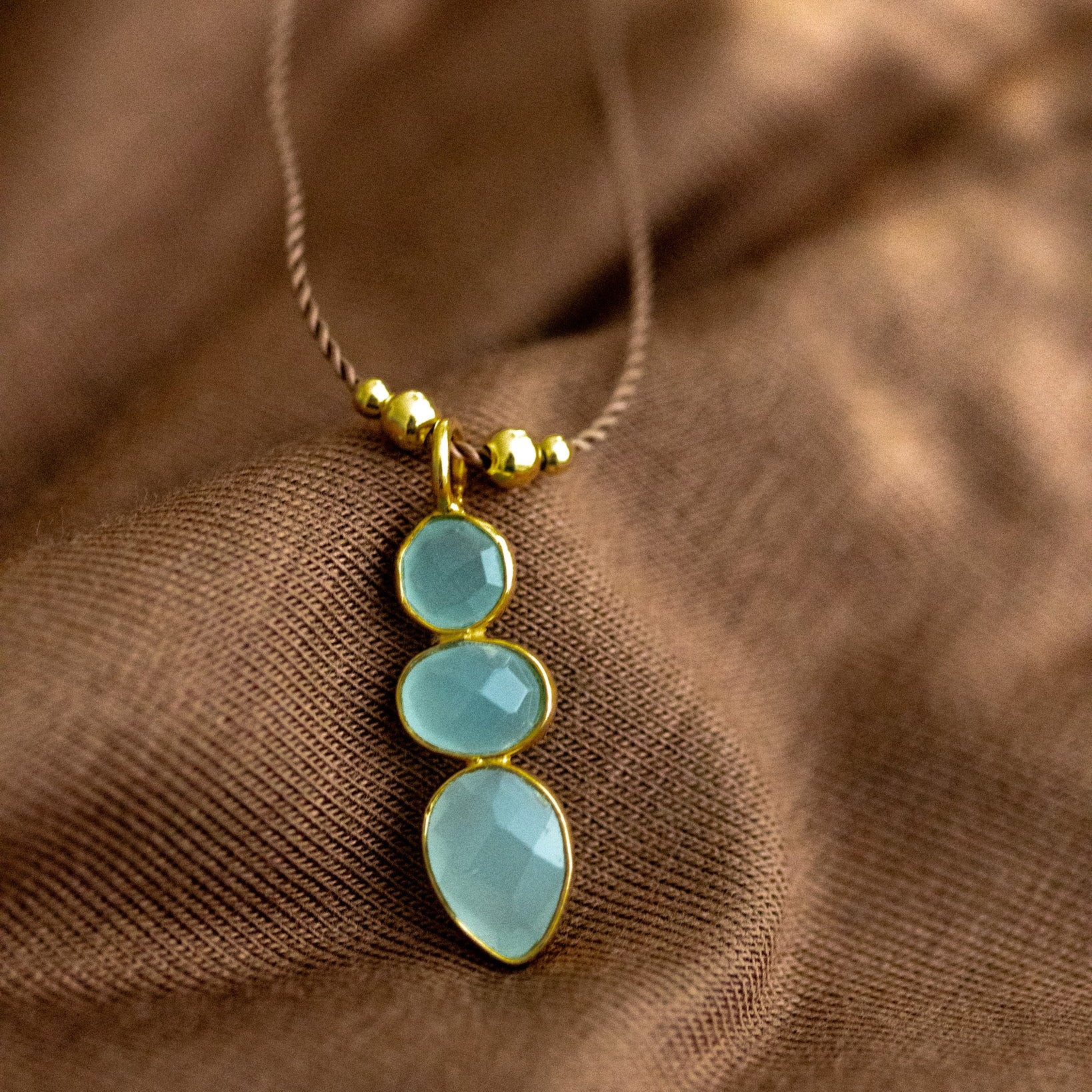 Eine lange Schnurkette mit einem länglichen Anhänger, der aus drei übereinander in gold gefassten Aqua Chalcedonen besteht, liegt auf braunem Stoff.