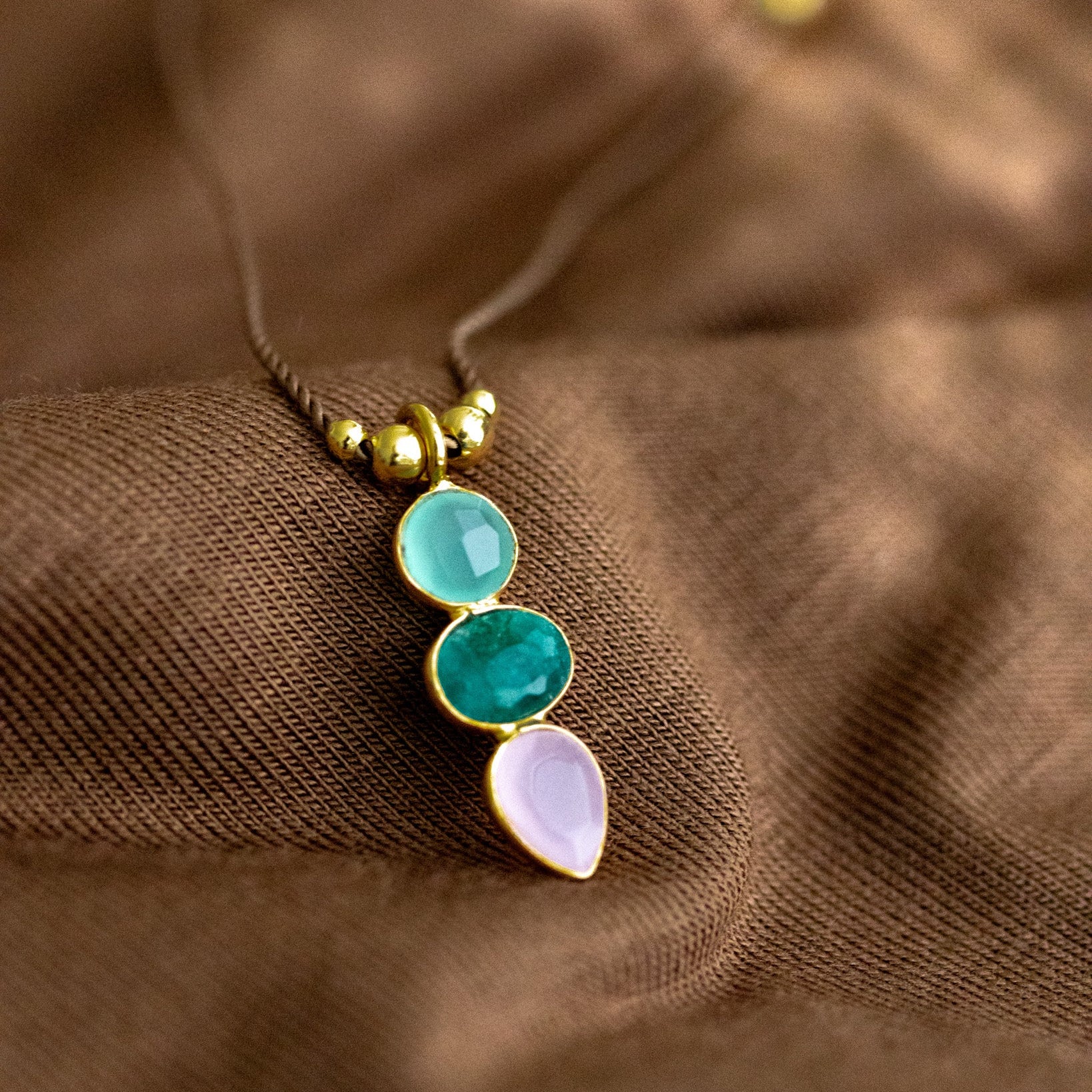 Eine dünne Schnurkette mit einem bunten Multi-Edelsteinanhänger und goldenen Zierelementen liegt auf braunem Hintergrund.