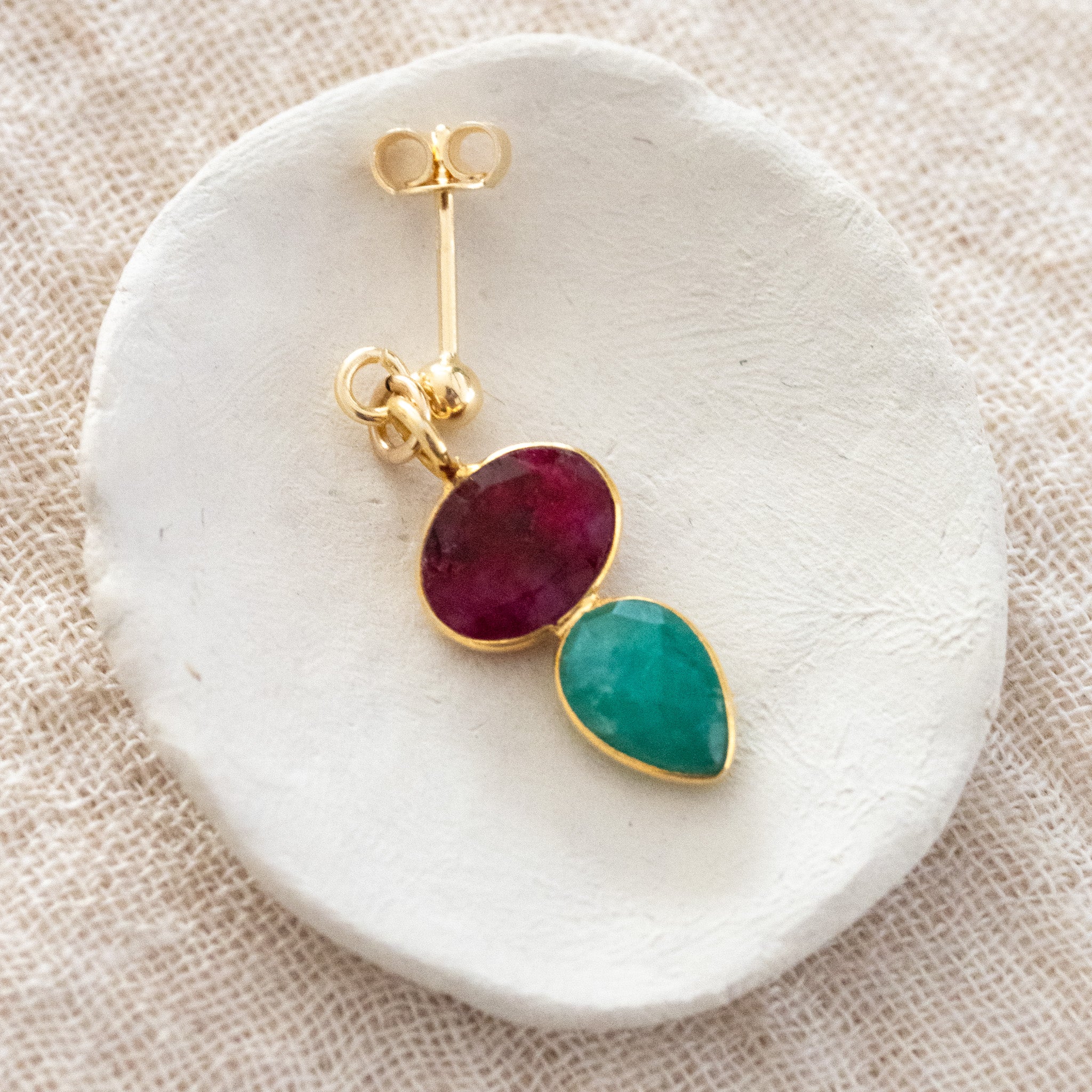 Ein Ohrstecker, der aus einem gold gerahmten Rubin und einem darunter angebrachten, ebenfalls gold gefassten, Smaragd besteht, liegt auf einem weißen Schälchen.