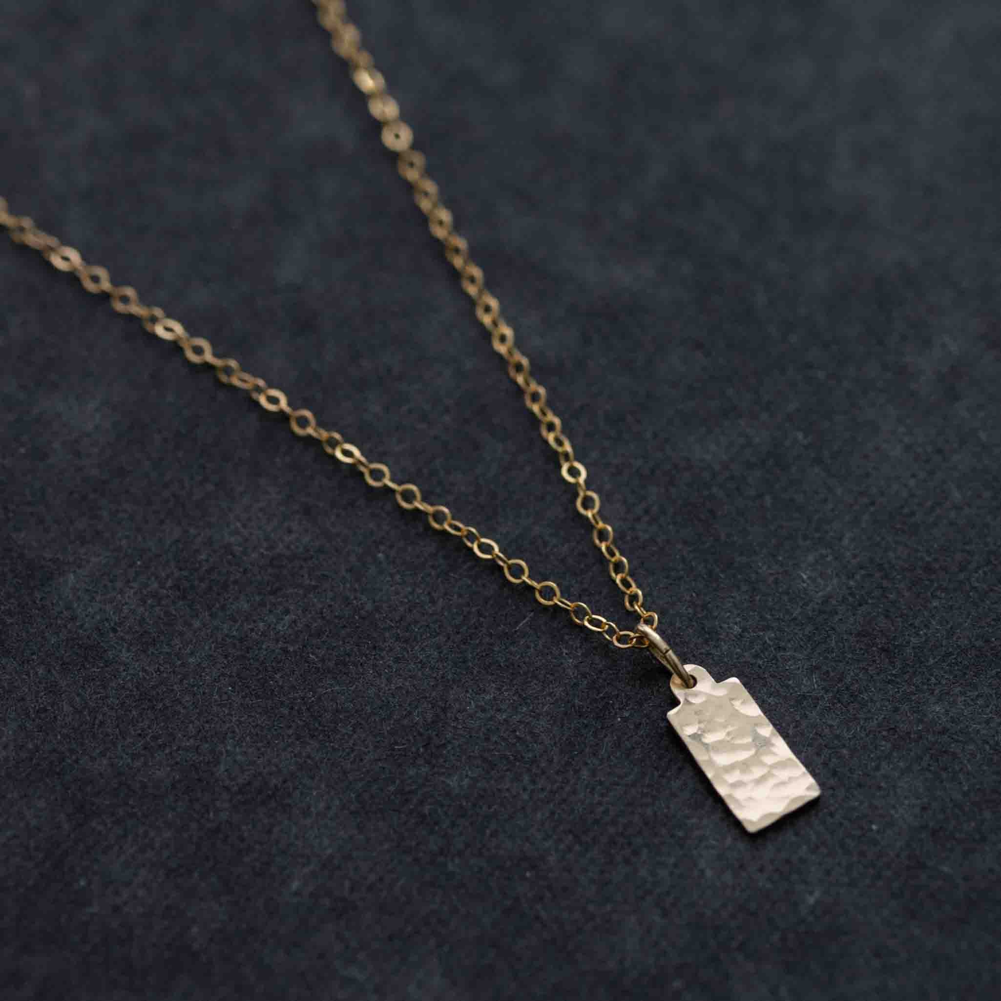 Eine feine und kurze Goldkette mit einem rechteckigen, gehämmerten Plättchen-Anhänger liegt auf schwarzem Hintergrund.