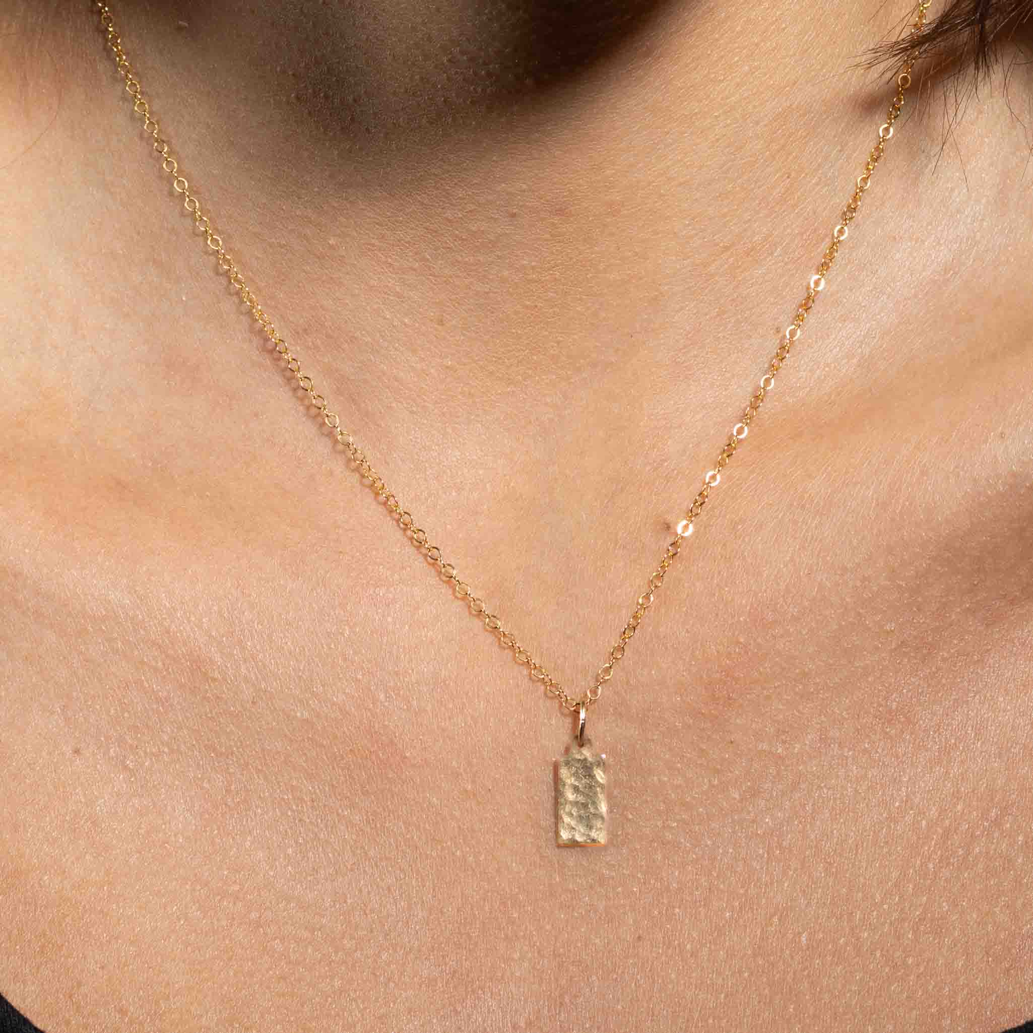 Eine feine und kurze Goldkette mit einem rechteckigen, gehämmerten Plättchen-Anhänger hängt vom Hals des Models herab.