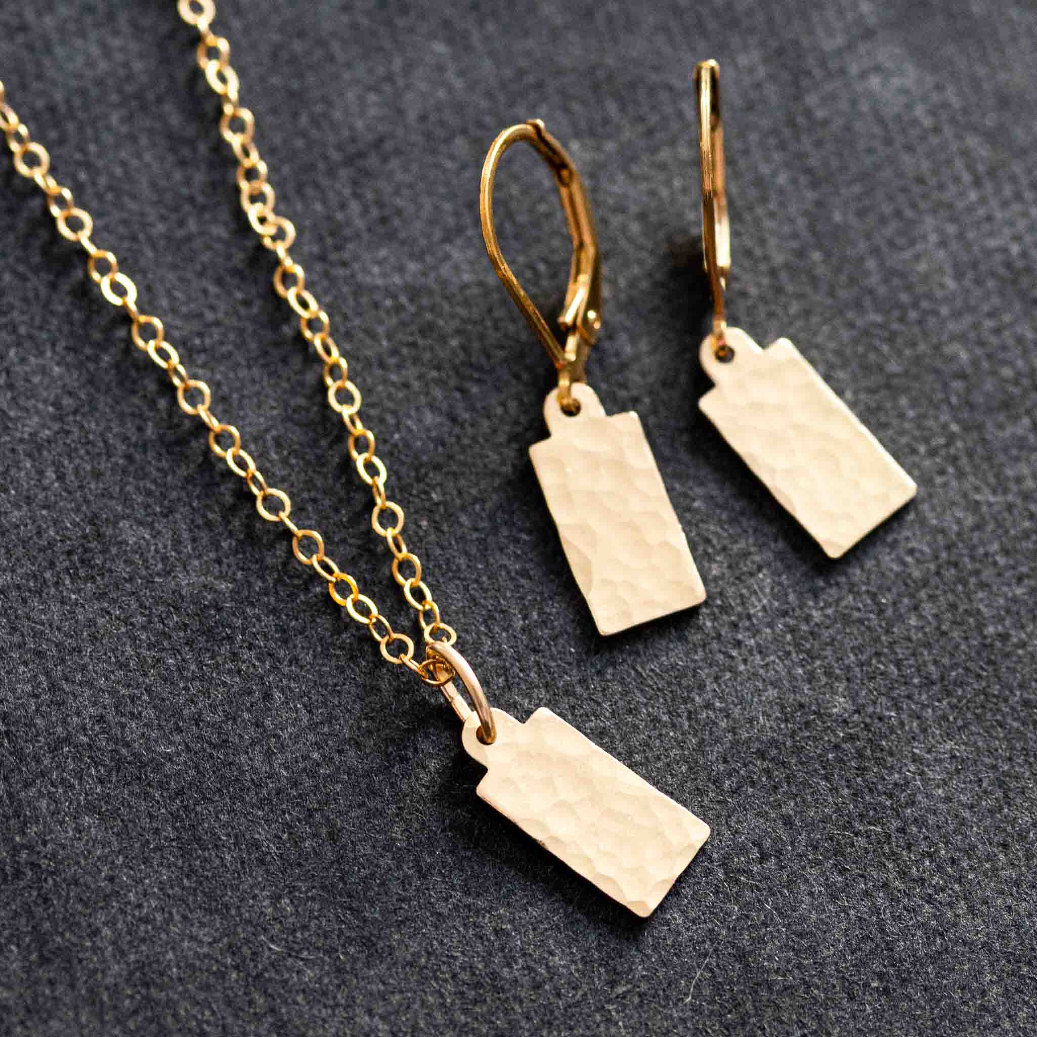 Ein Schmuckset liegt auf schwarzem Hintergrund. Es besteht aus einer feinen Goldkette mit rechteckigem Plättchen-Anhänger und passenden Ohrringen.
