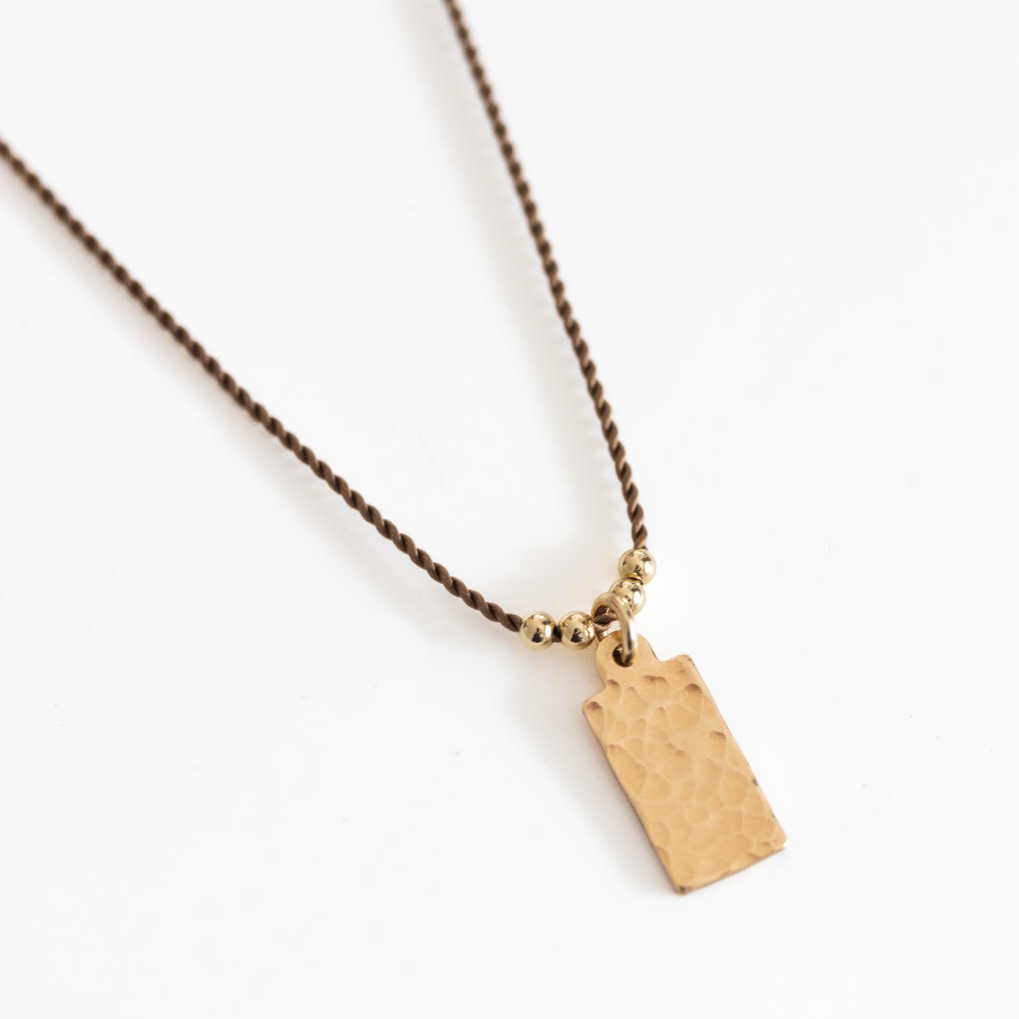 Eine feine, beige Kordelkette mit einem rechteckigen, gehämmerten Plättchenanhänger auf weißem Hintergrund.