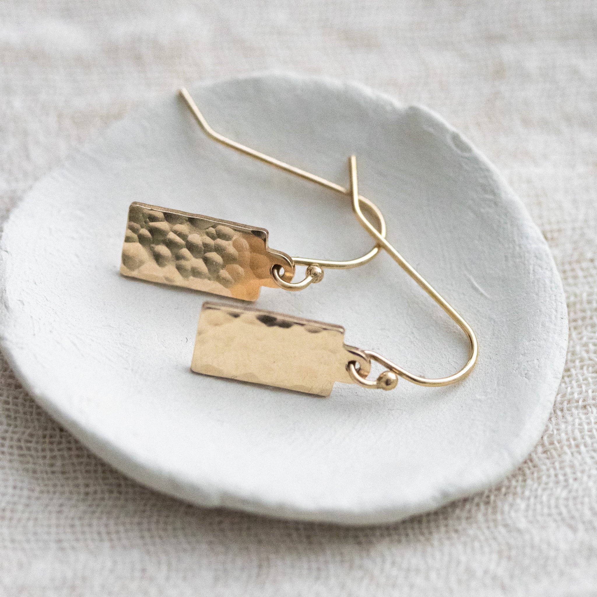 Auf einem kleinen, weißen Schälchen liegt ein Paar goldener Ohrringe mit einem rechteckigen, gehämmerten Plättchen an einfachen Ohrhaken.