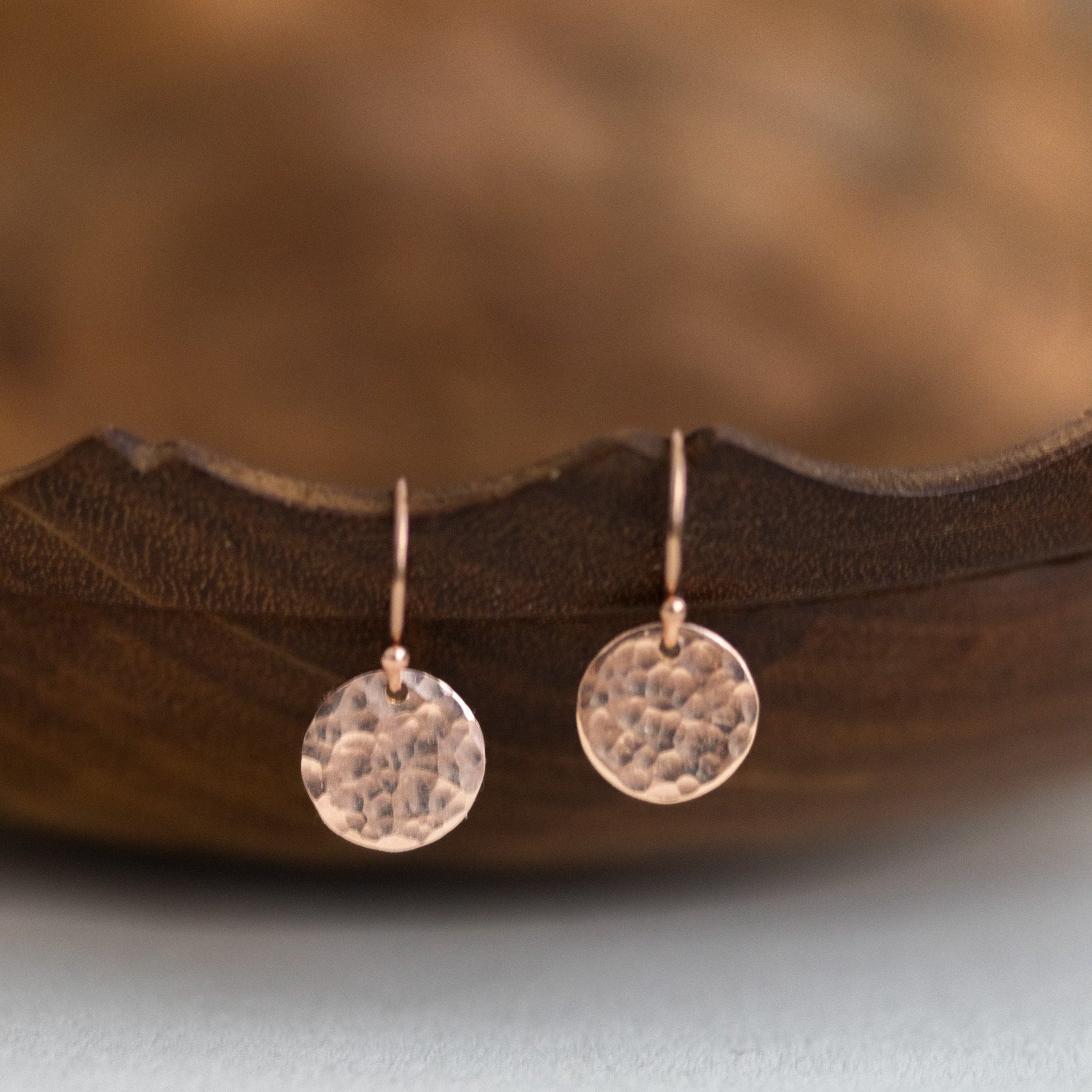 Ein Paar rosé goldener Ohrringe mit gehämmerten Plättchen hängt an einer gewellten, braunen Schale.