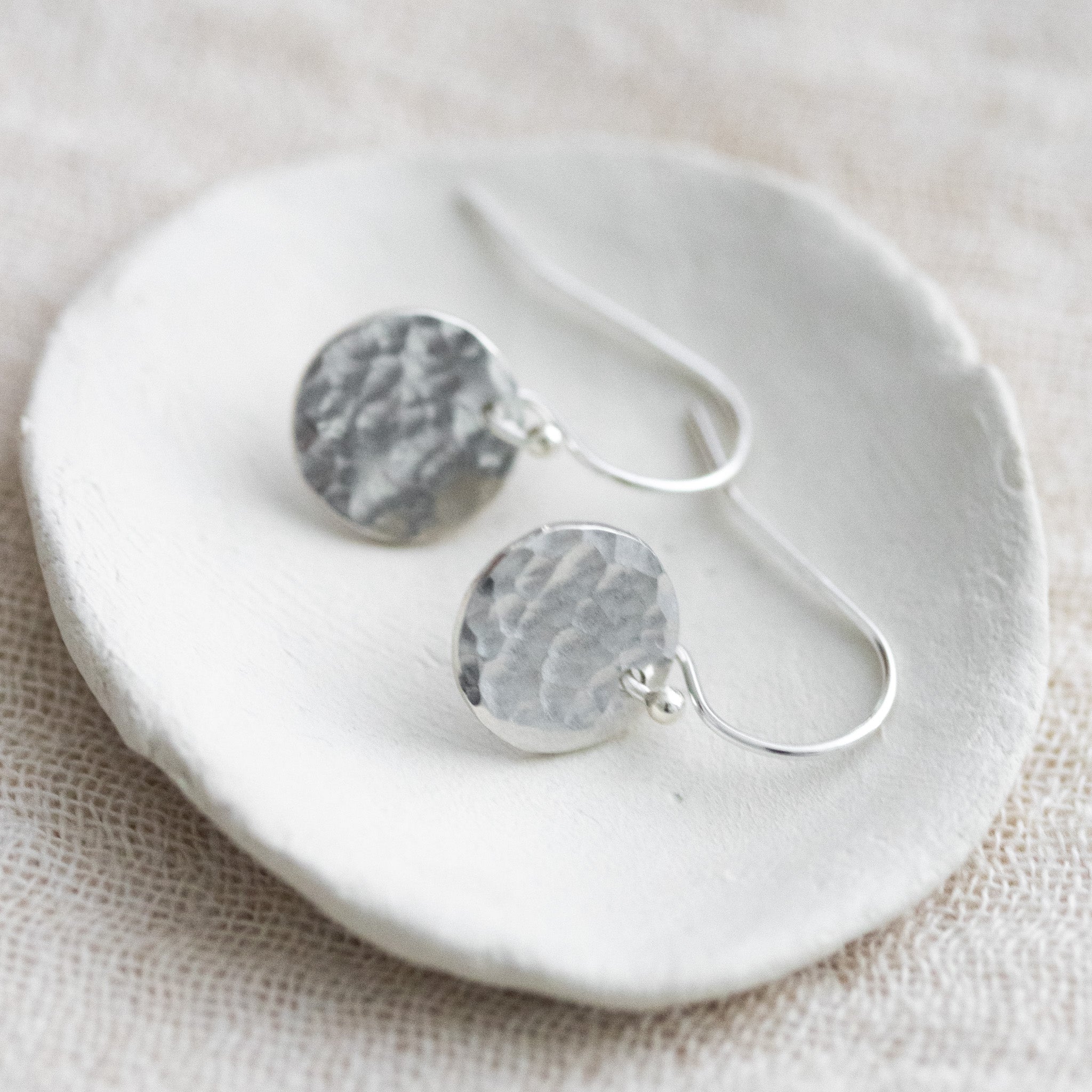Auf einem weißen Schälchen liegt ein Paar Silber-Ohrringe mit jeweils einem gehämmerten, runden Plättchen an einem einfachen Ohrhaken.