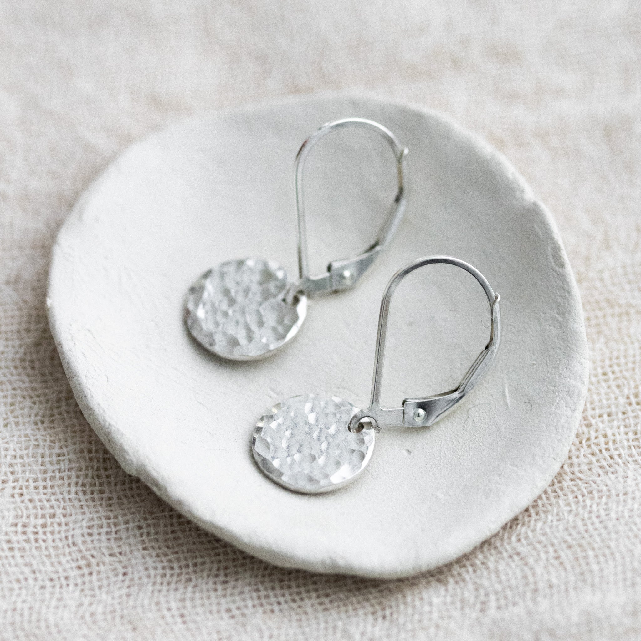 Auf einem kleinen, weißen Schälchen liegt ein Paar 925 Silber-Ohrringe mit jeweils einem runden, gehämmerten Plättchen an einer Klappbrisur.