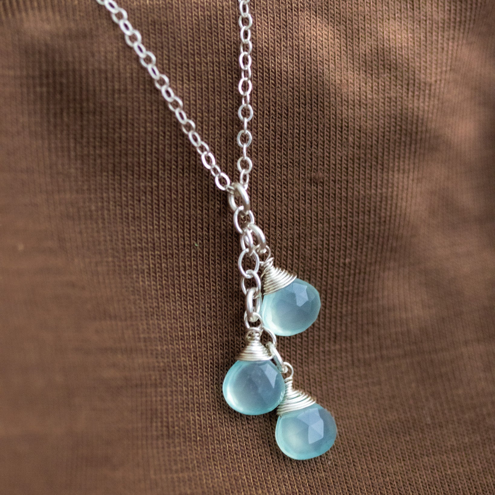 Eine feine Silberkette mit drei untereinander angebrachten Aqua Chalzedon-Tropfen lehnt an einem braunen Hintergrund.