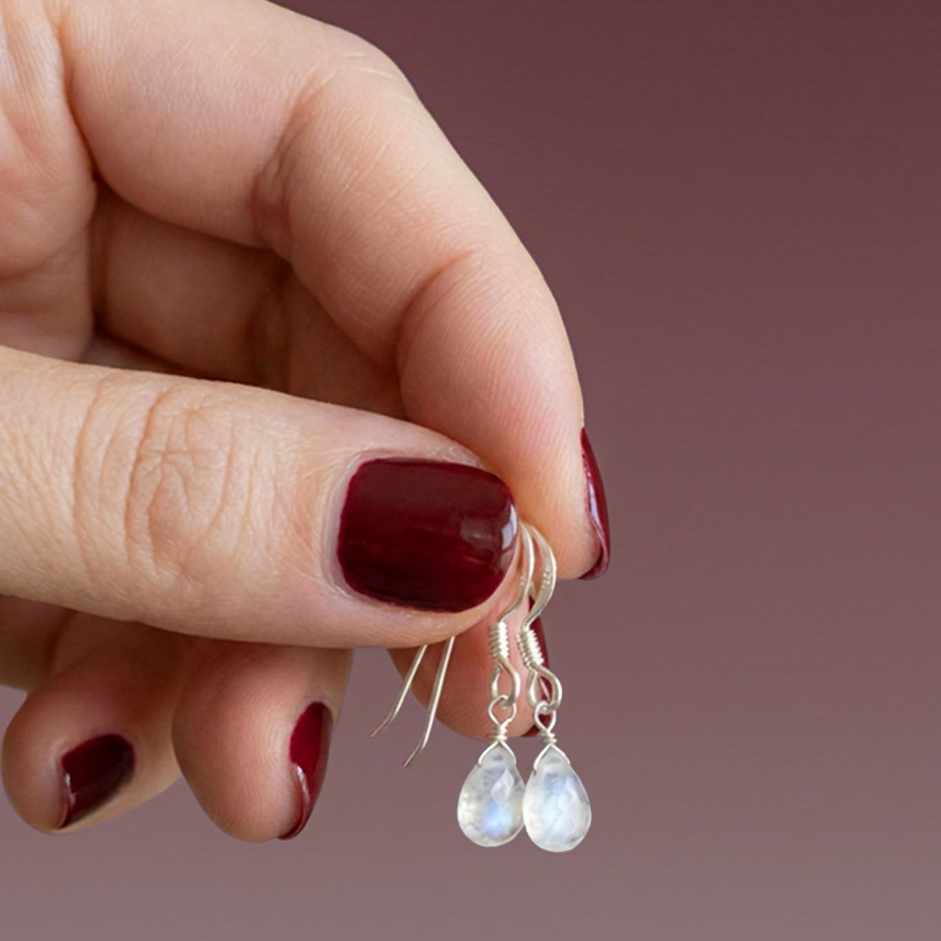 Eine Hand mit rot lackierten Fingernägeln hält ein Paar Silber-Ohrringe mit Mondstein-Tropfen.