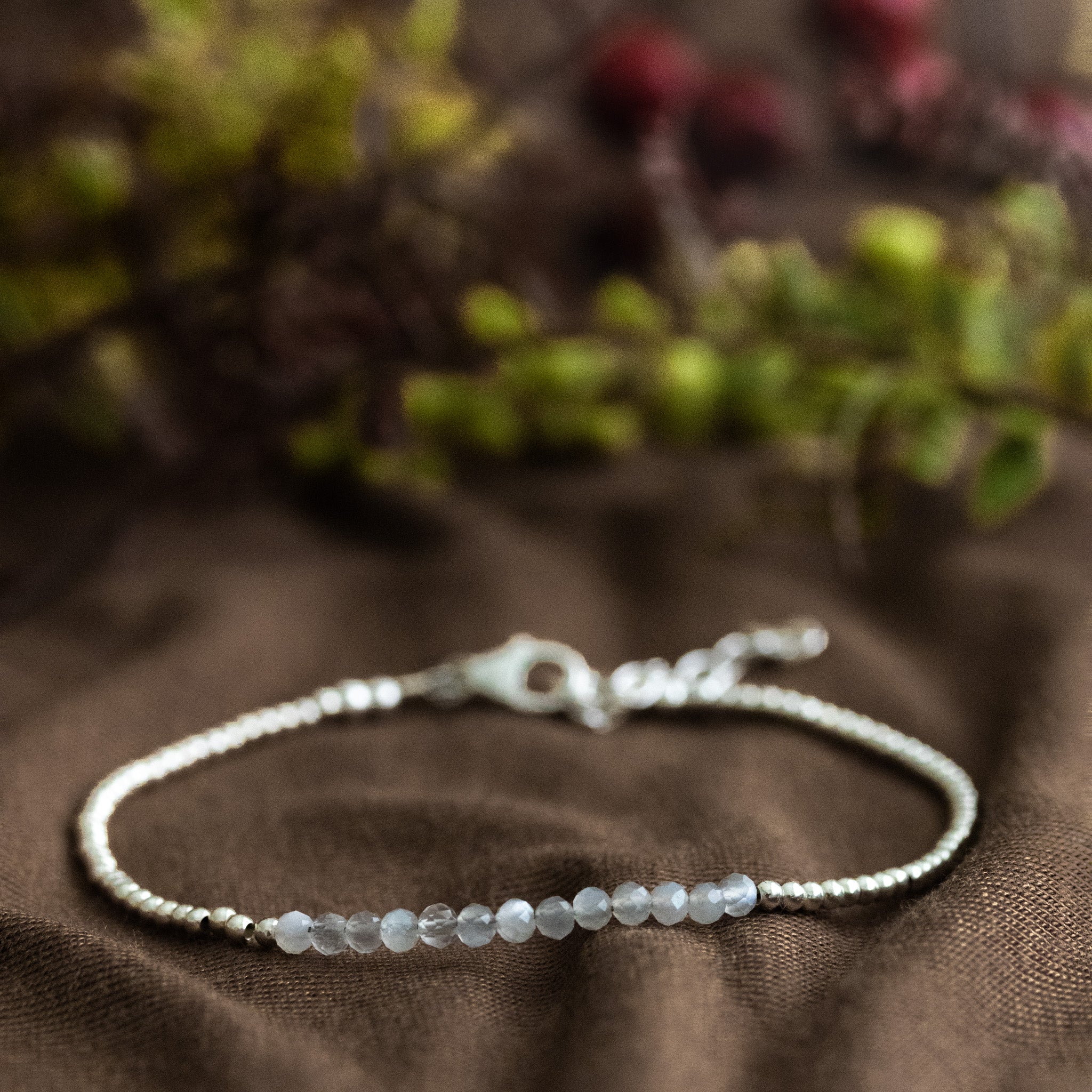 Ein ultra dünnes Silberarmband mit grauen Mondsteinperlen in der Mitte liegt auf braunem Stoff. Verschwommener floraler Hintergrund.