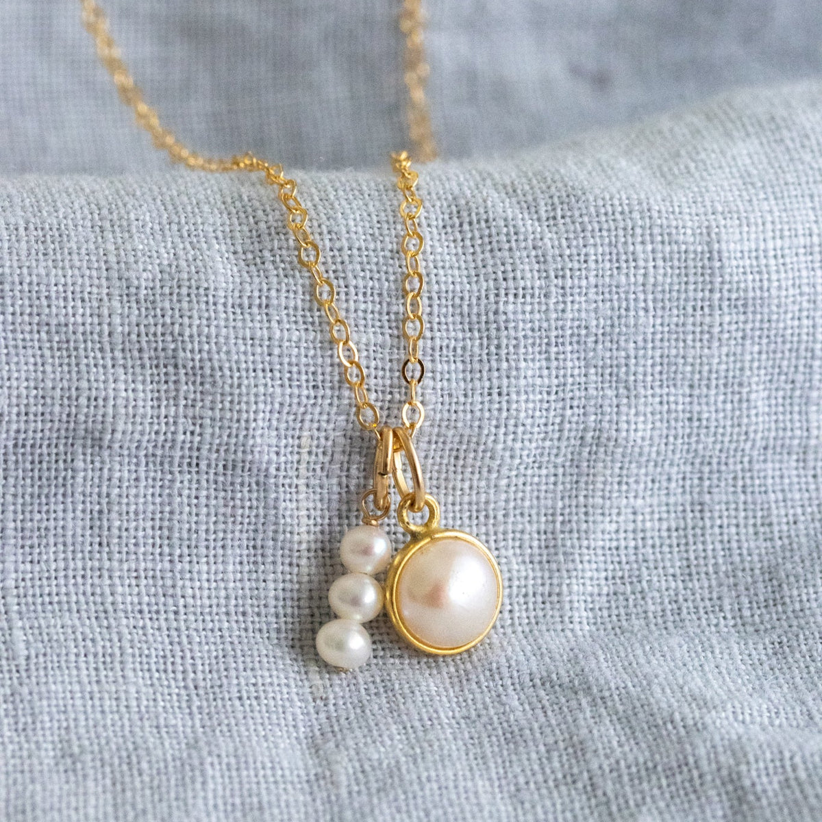 Eine feine Goldkette mit zwei unterschiedlichen Perlen-Anhängern angelehnt an grauen Stoff.