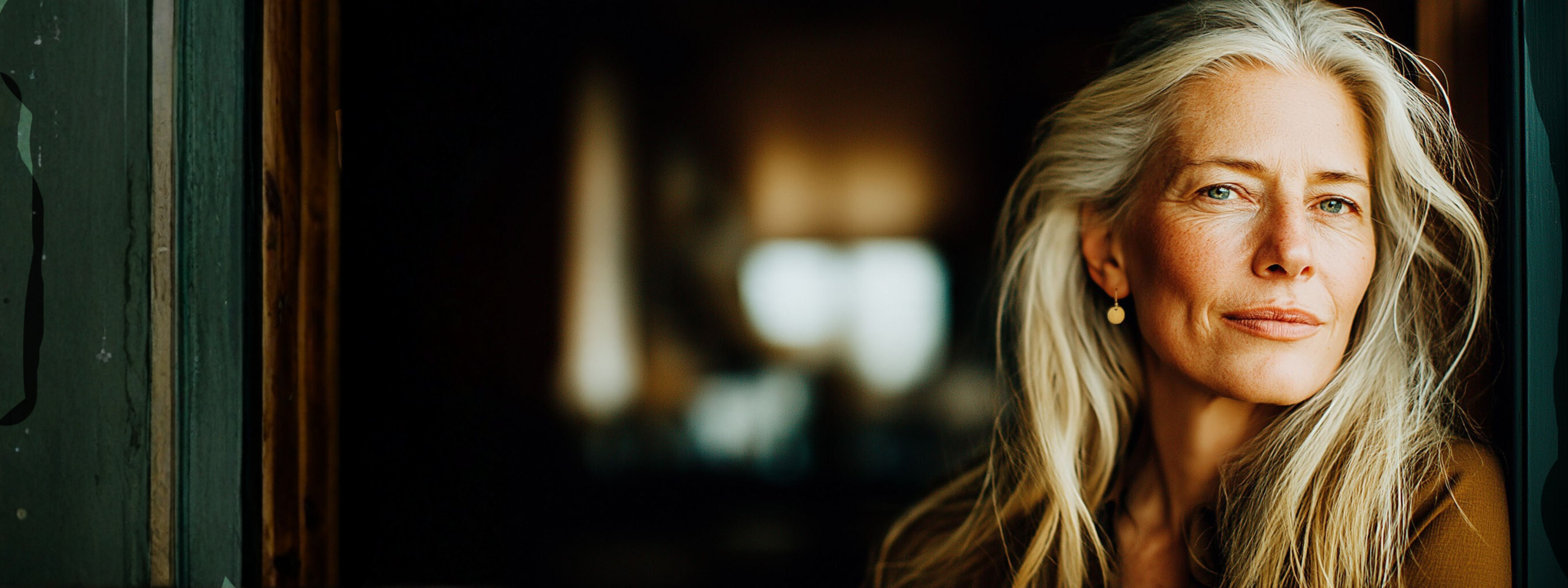 Eine schöne, mittelalte Frau mit langen, grauen Haaren und einem goldenen Ohrring steht in einem dunklen Raum an die Wand gelehnt und schaut in die Kamera.