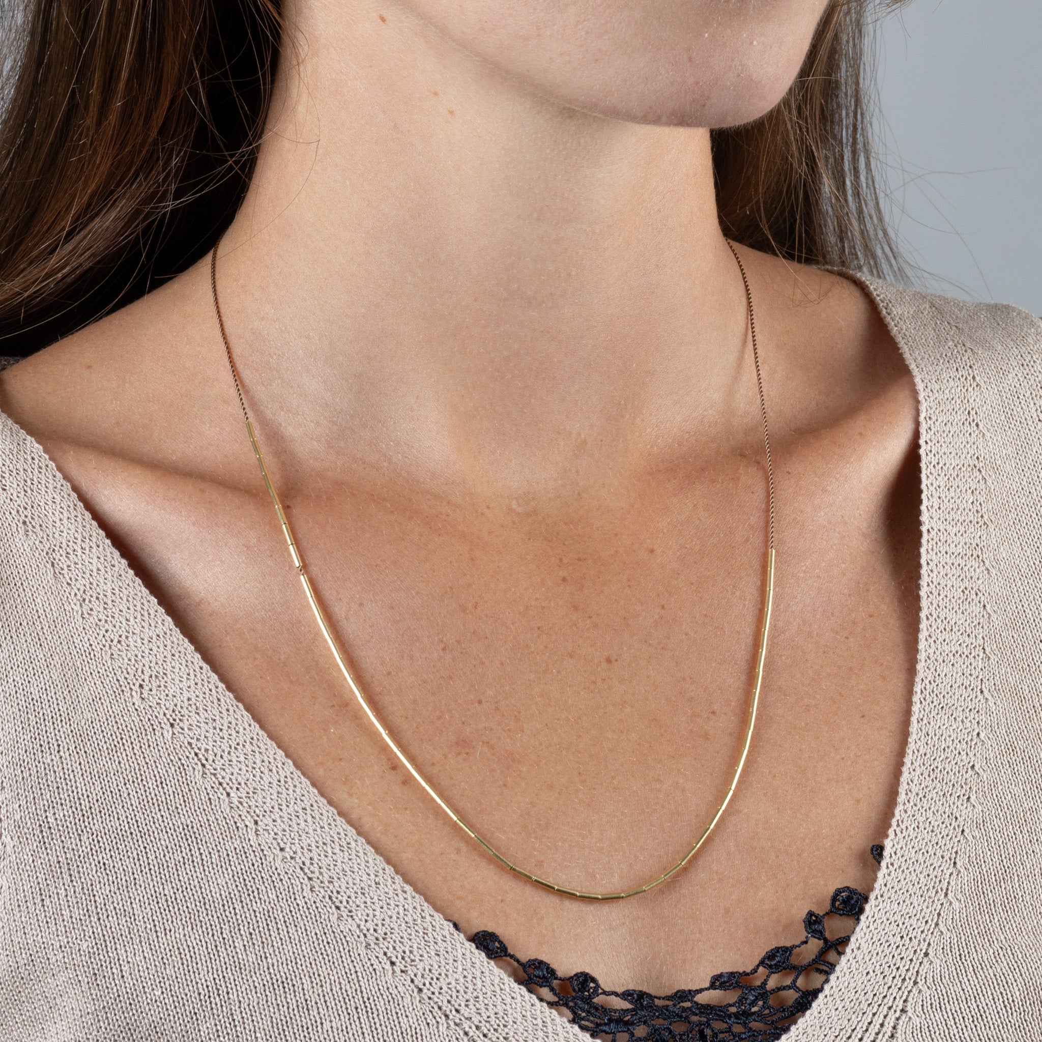 Eine zierliche Schnurkette mit einer fließenden Linie aus schmalen Goldröhrchen, getragen von einer Frau in einem beigefarbenen V-Ausschnitt-Pullover.