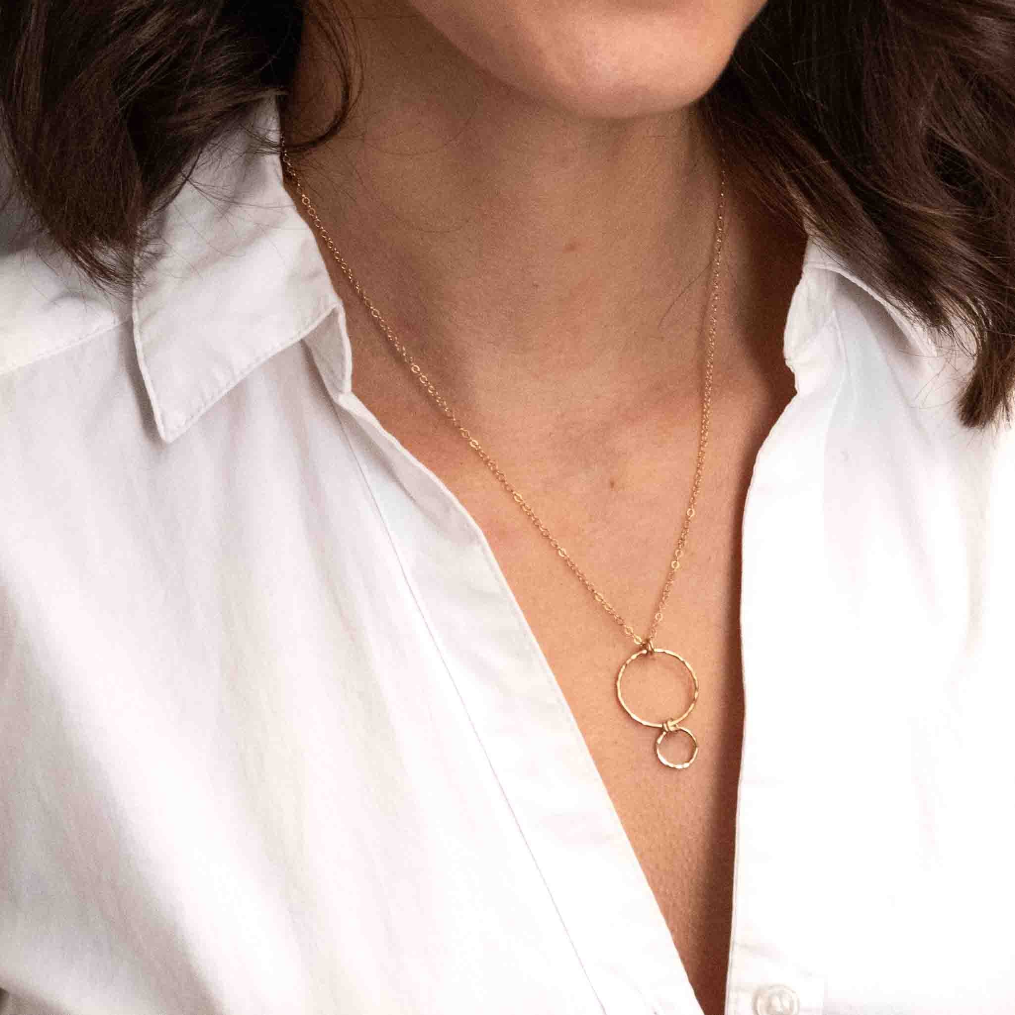 Eine feine Goldkette mit einem Anhänger, bestehend aus einem größeren Ring, an dem ein kleinerer Ring hängt, hängt vom Hals des Models.