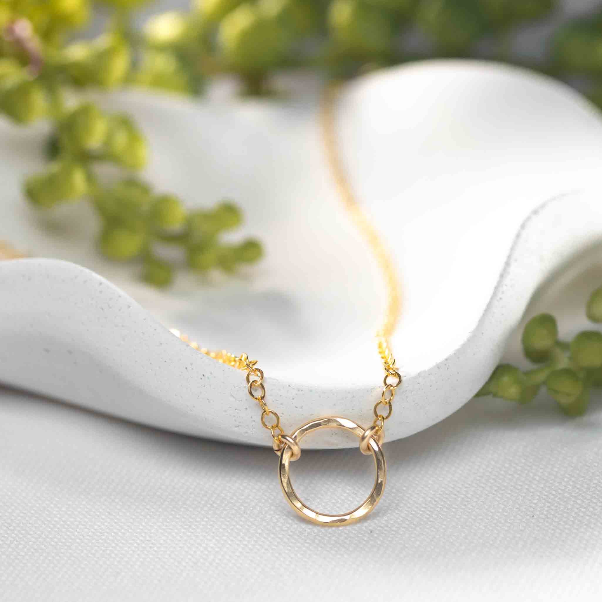 Eine feine Goldkette mit offenem Ring-Anhänger lehnt an einem weißen Schälchen, grüne Blätter im Hintergrund.