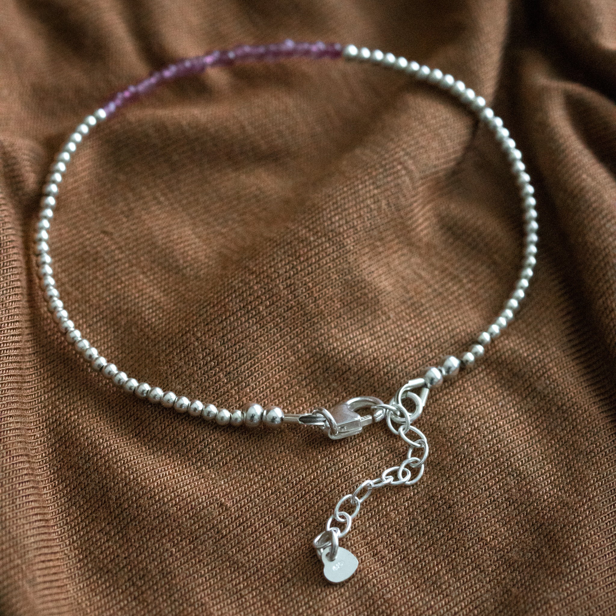 Ein sehr dünnes Silberarmband mit winzigen rosa Turmalinen in der Mitte liegt auf braunem Stoff.