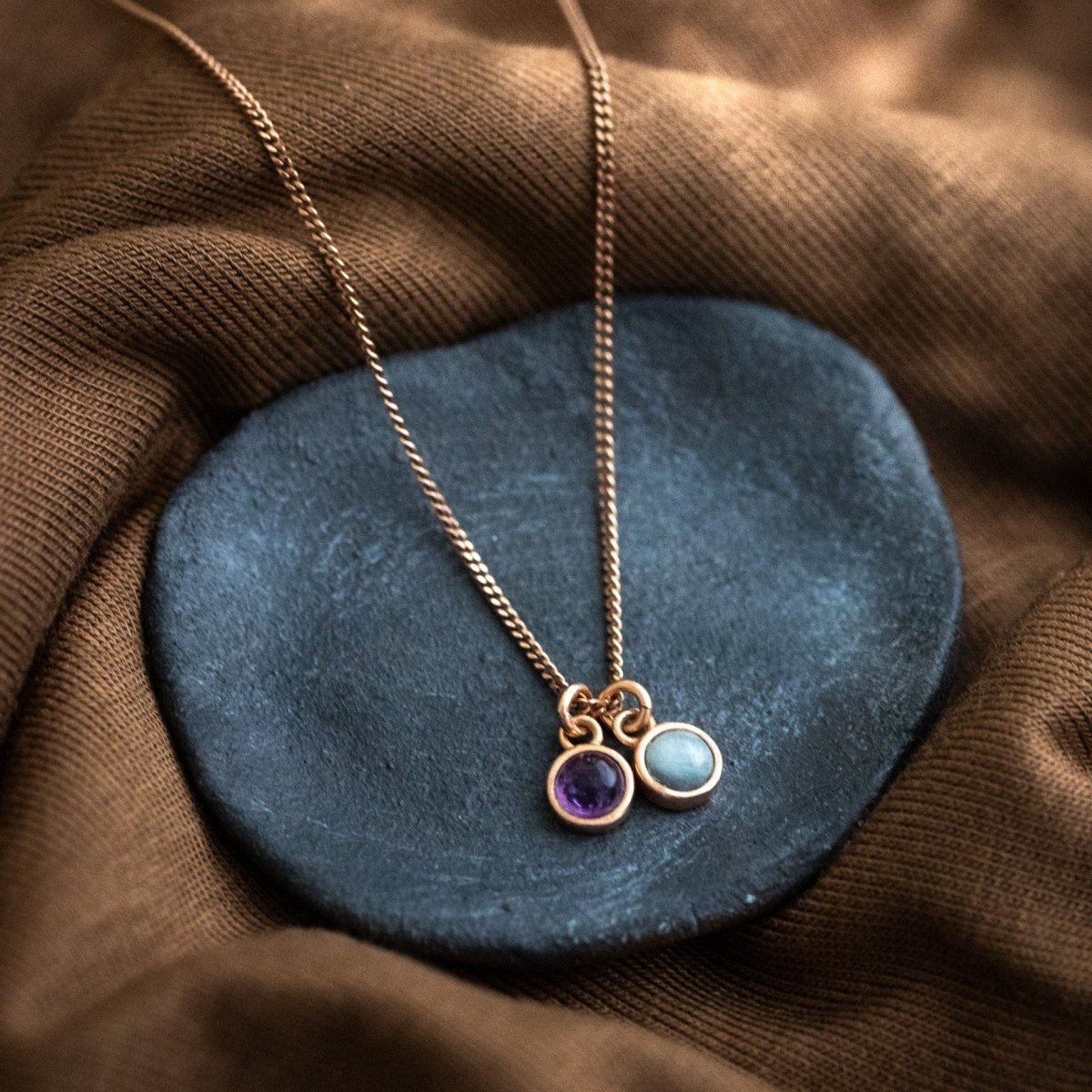 Eine feine rosé vergoldete Halskette mit zwei winzigen, runden Anhängern, einem Amethyst und einem Aquamarin, liegt auf einem dunkelgrauen Steinschälchen.