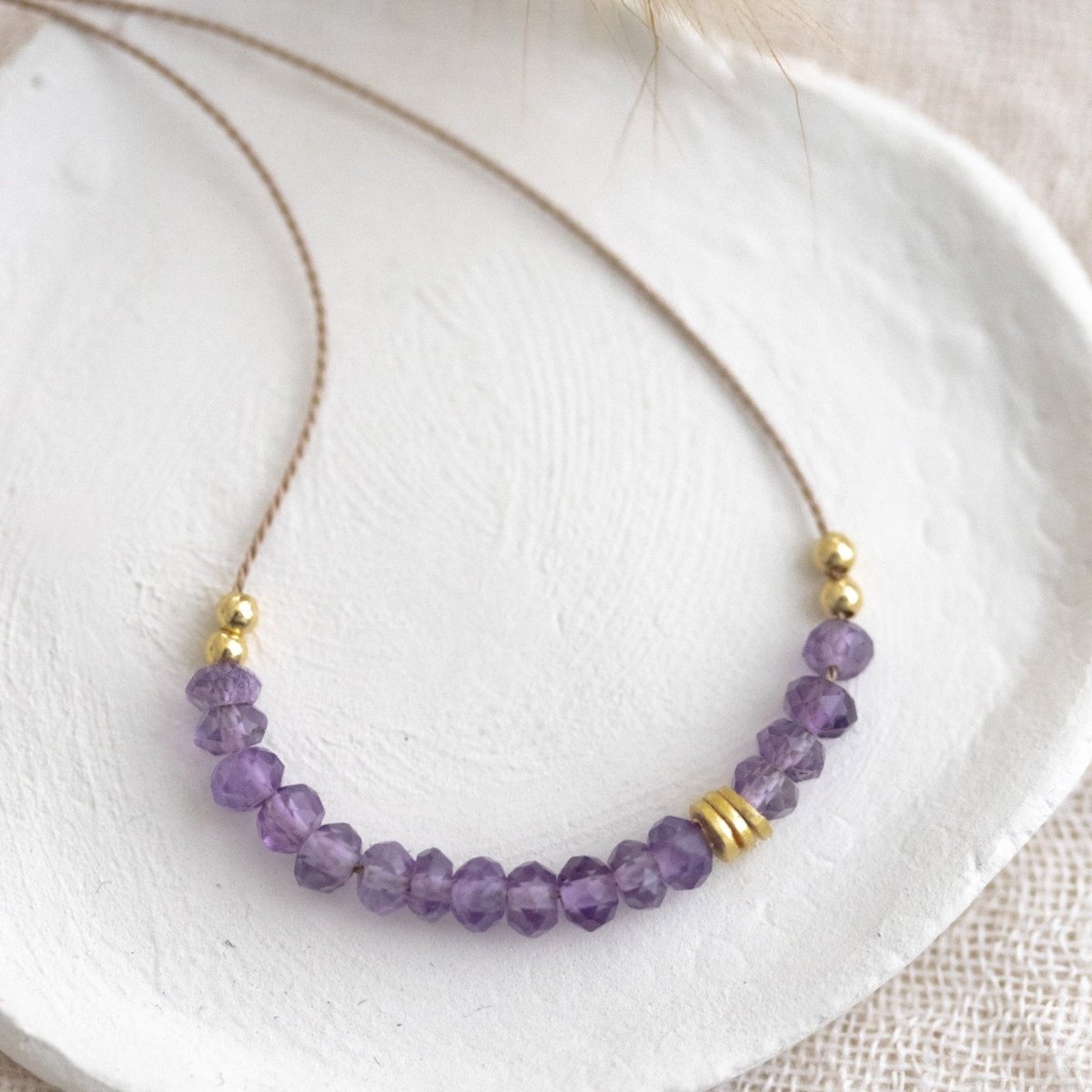 Auf einem weißen Schälchen liegt eine kurze Halskette. Sie besteht aus einer dünnen Seidenschnur mit einer Reihe kleiner, violetter Amethyst-Rondellen und goldenen Zierperlen in der Mitte.