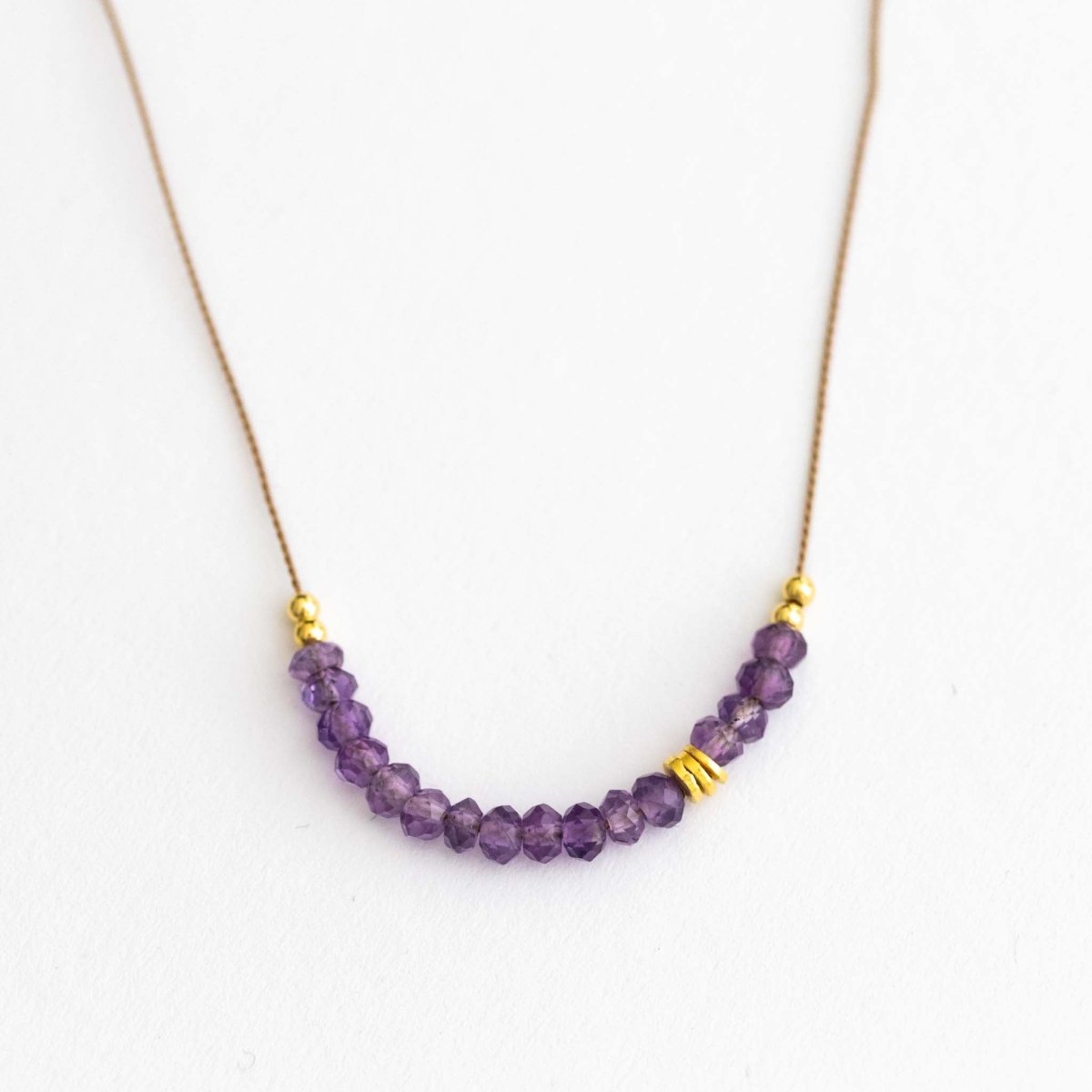 Auf weißen Hintergrund liegt eine kurze Halskette. Sie besteht aus einer dünnen Seidenschnur mit einer Reihe kleiner, violetter Amethyst-Rondellen und goldenen Zierperlen in der Mitte.