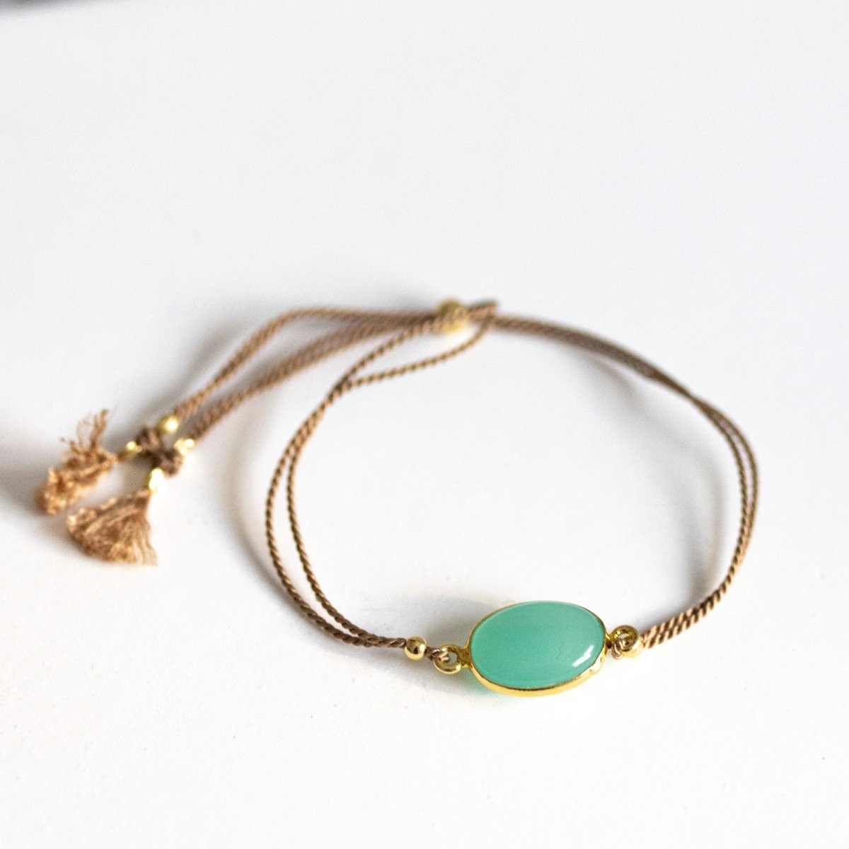 Ein beiges Seidenarmband mit einem gold gefassten, ovalen Aqua Chalcedon liegt auf weißem Hintergrund.