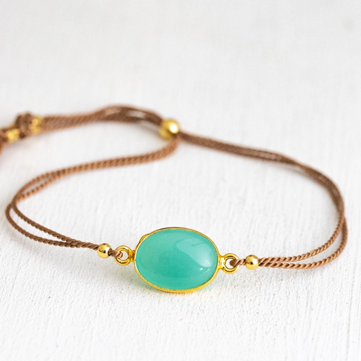 Ein beiges Seidenarmband mit einem gold gefassten, ovalen Aqua Chalcedon liegt auf weißem Hintergrund.