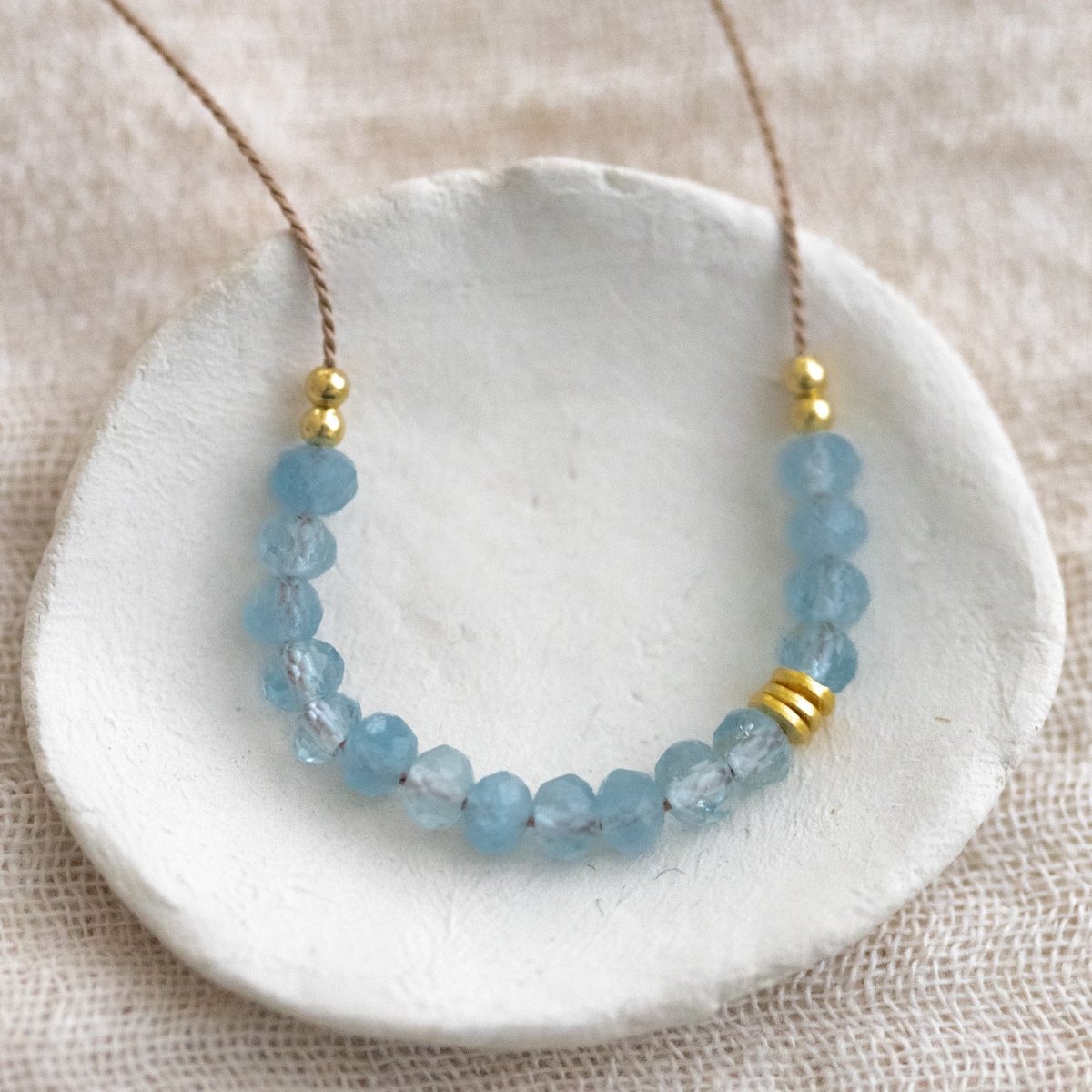 Auf einem weißen Schälchen liegt eine Halskette, die aus dünner, beiger Seidenschnur besteht, auf die in der Mitte eine Reihe kleiner Aquamarin-Rondelle gezogen wurden.