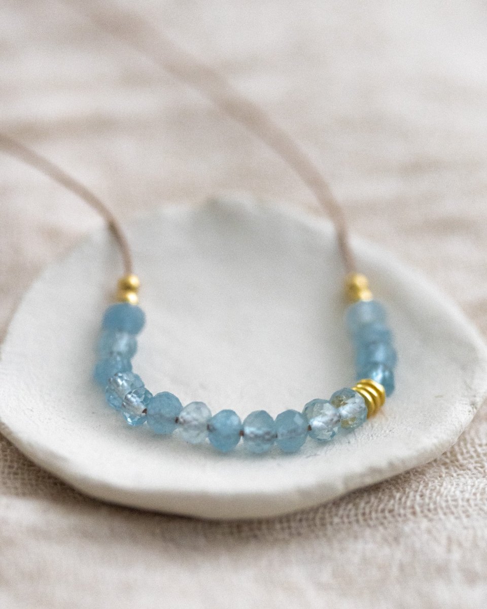 Auf einem weißen Schälchen liegt eine Halskette, die aus dünner, beiger Seidenschnur besteht, auf die in der Mitte eine Reihe kleiner Aquamarin-Rondelle gezogen wurden.