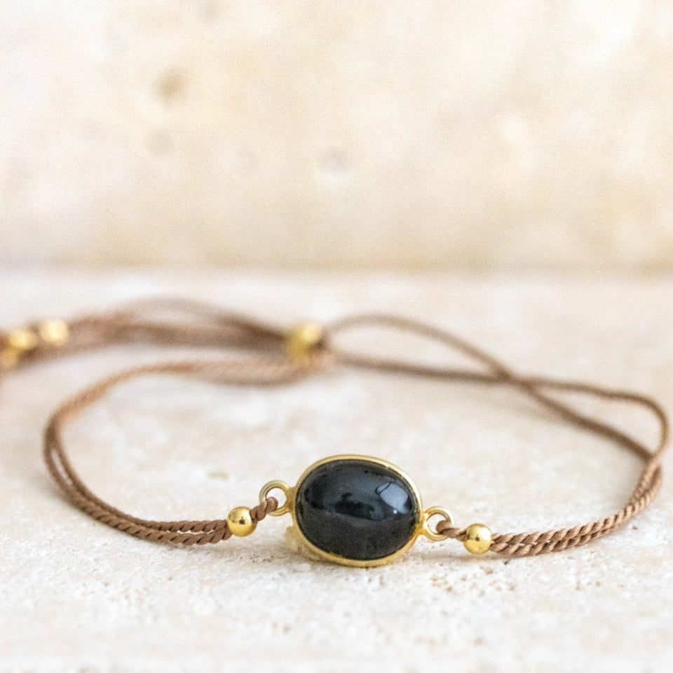 Ein beiges Seidenarmband mit einem Black Star Edelstein in der Mitte auf einer hellen Steinplatte.