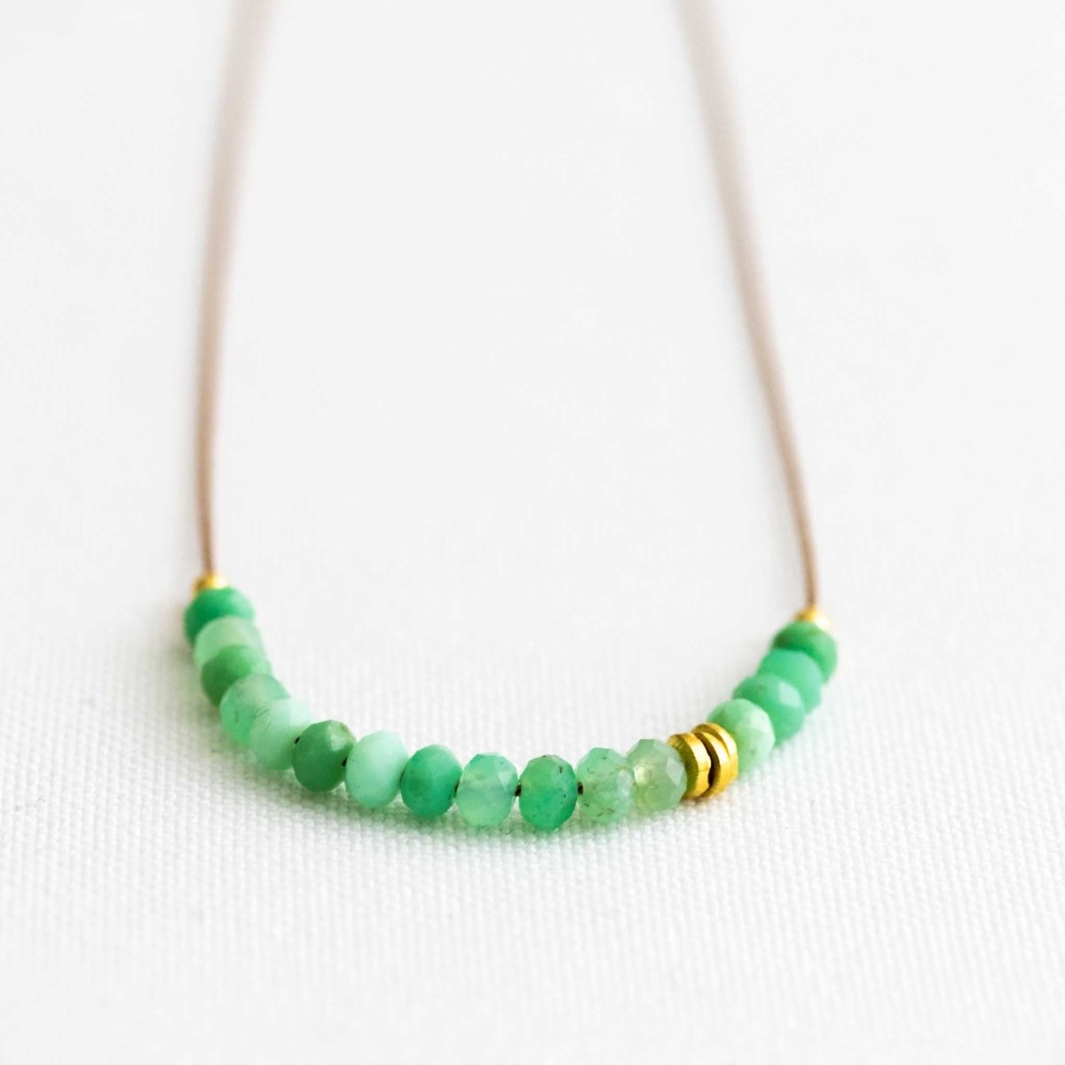 Auf weißem Hintergrund liegt eine dünne Halskette. Sie besteht aus einer beigen Seidenschnur, auf die eine Reihe grüner Chrysopras-Rondelle und einige goldene Zierperlen aufgezogen sind.