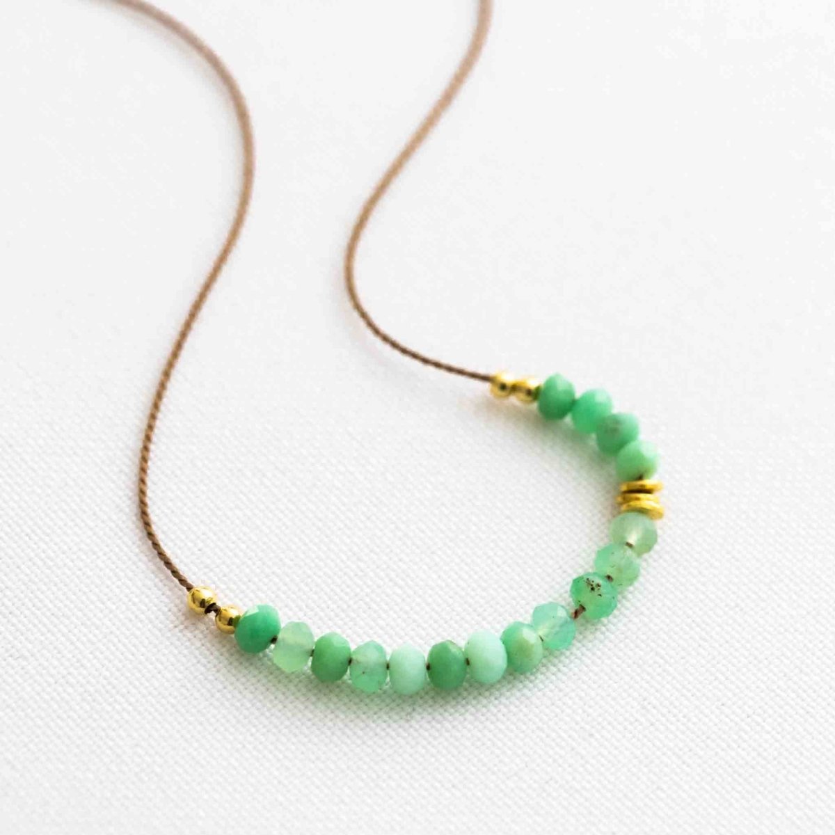 Auf weißem Hintergrund liegt eine dünne Halskette. Sie besteht aus einer beigen Seidenschnur, auf die eine Reihe grüner Chrysopras-Rondelle und einige goldene Zierperlen aufgezogen sind.
