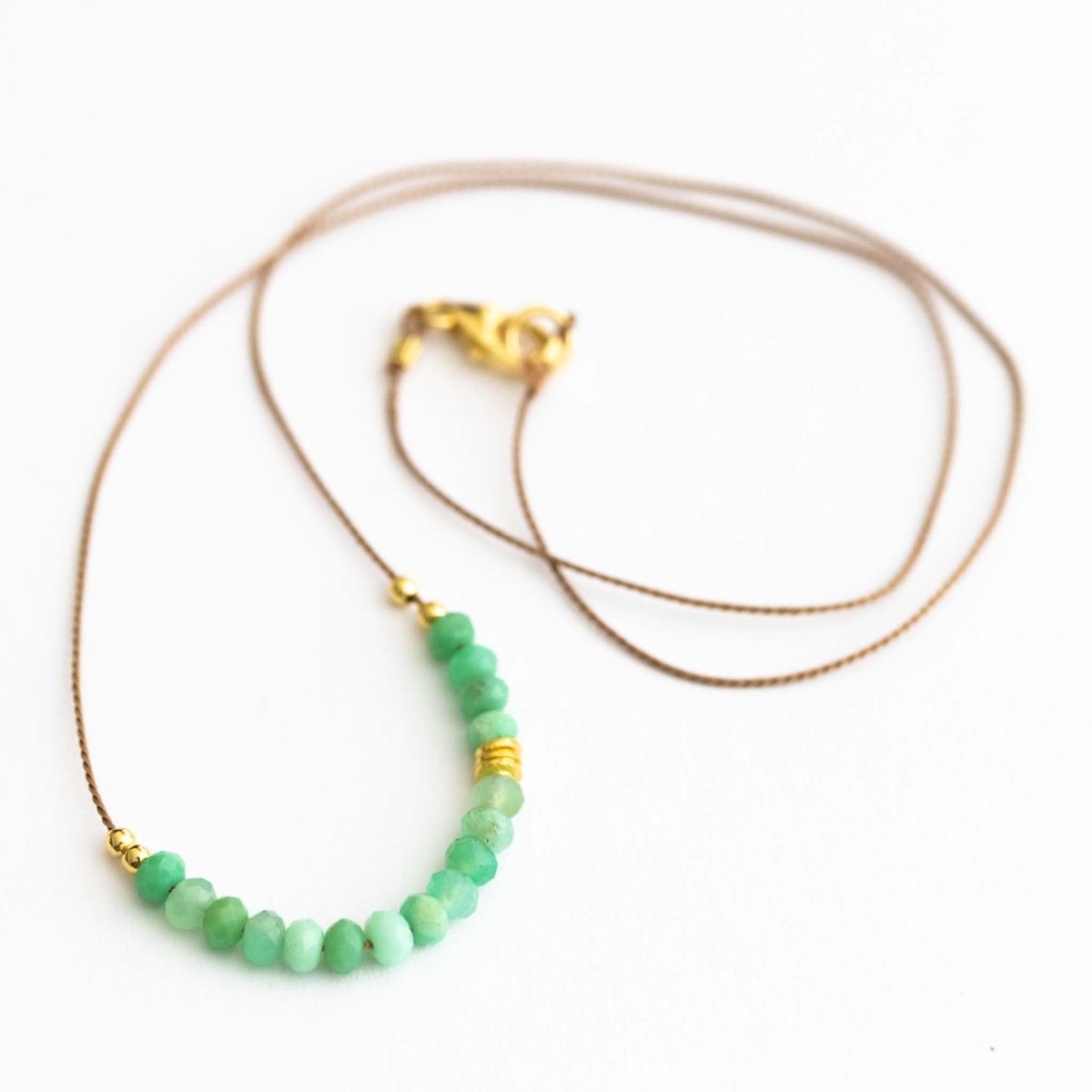 Auf weißem Hintergrund liegt eine dünne Halskette. Sie besteht aus einer beigen Seidenschnur, auf die eine Reihe grüner Chrysopras-Rondelle und einige goldene Zierperlen aufgezogen sind.