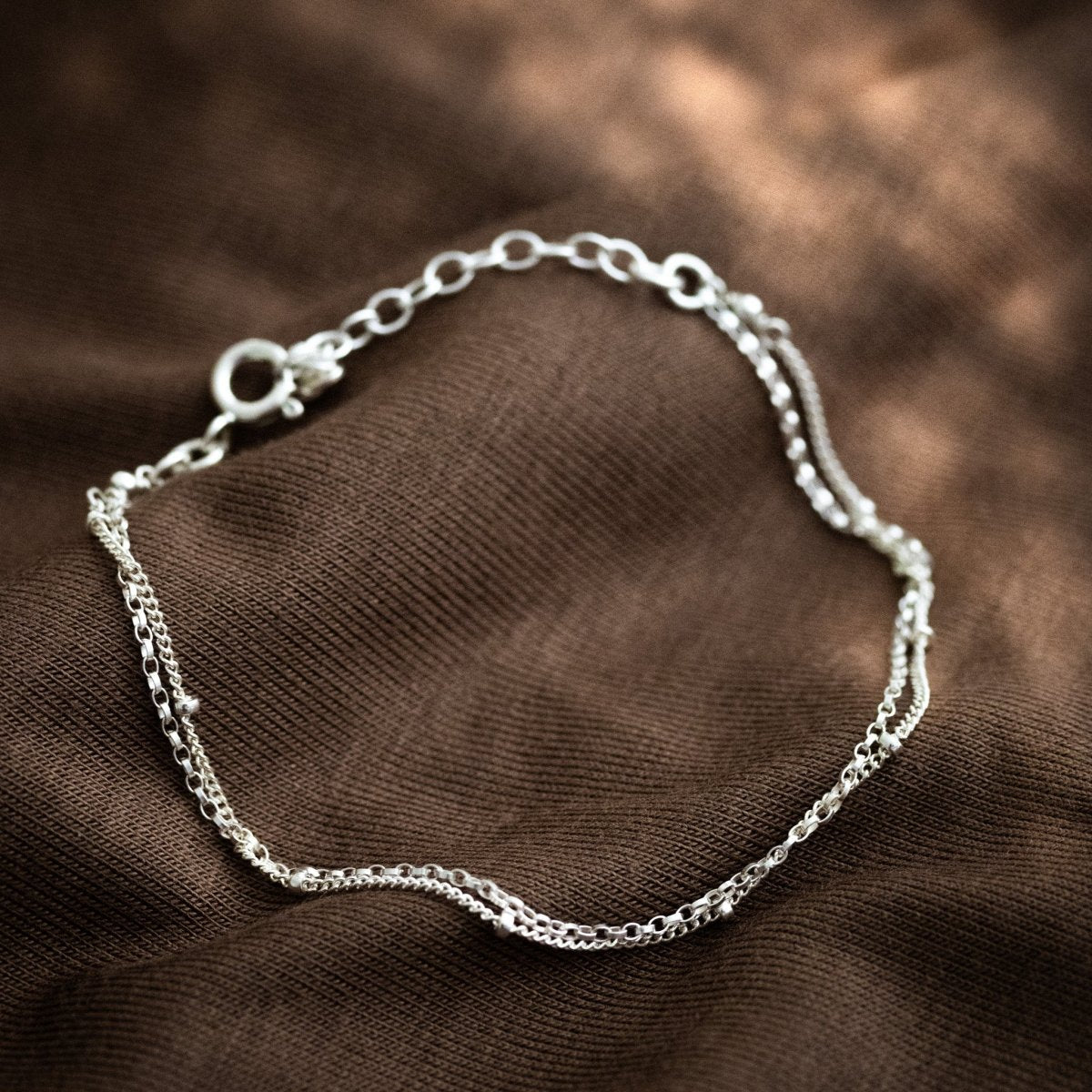 Ein doppelreihiges Silber-Armband liegt auf braunem Stoff.