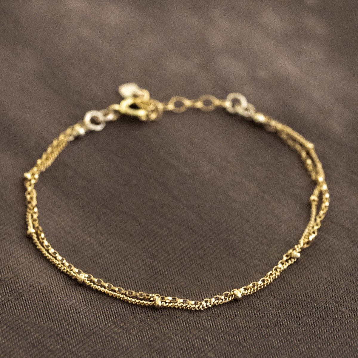 Auf braunem Hintergrund liegt ein zierliches, doppelt gelegtes Armband aus Satellitenkette und Rolokette, beide in gold.