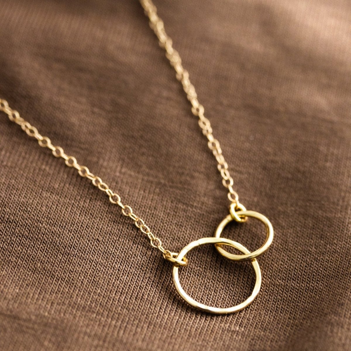 Eine feine Goldkette mit zwei ineinander verschlungenen Ringen in der Mitte liegt auf einem braunen Hintergrundstoff.