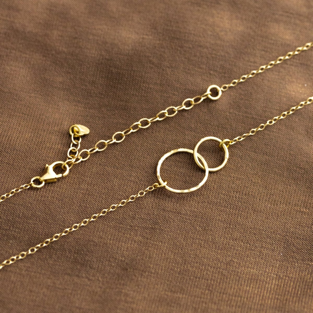 Eine feine Goldkette mit zwei ineinander verschlungenen Ringen und einem Karabinerverschluss und Verlängerungskettchen liegt auf braunem Hintergrund.