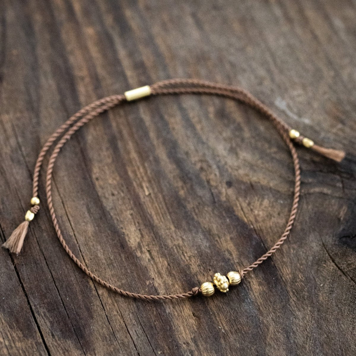 Ein dünnes, braunes Schnurarmband mit drei unterschiedlichen, goldenen Zierperlen in der Mitte auf Holz ausgelegt.