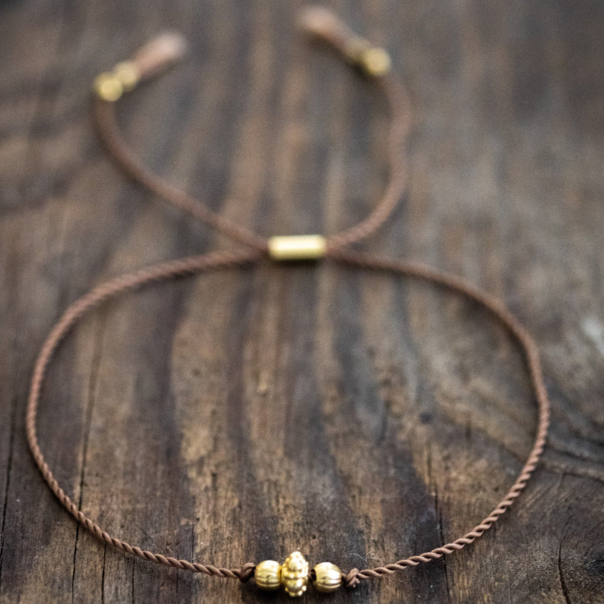 Ein dünnes, braunes Schnurarmband mit drei unterschiedlichen, goldenen Zierperlen in der Mitte auf Holz ausgelegt.