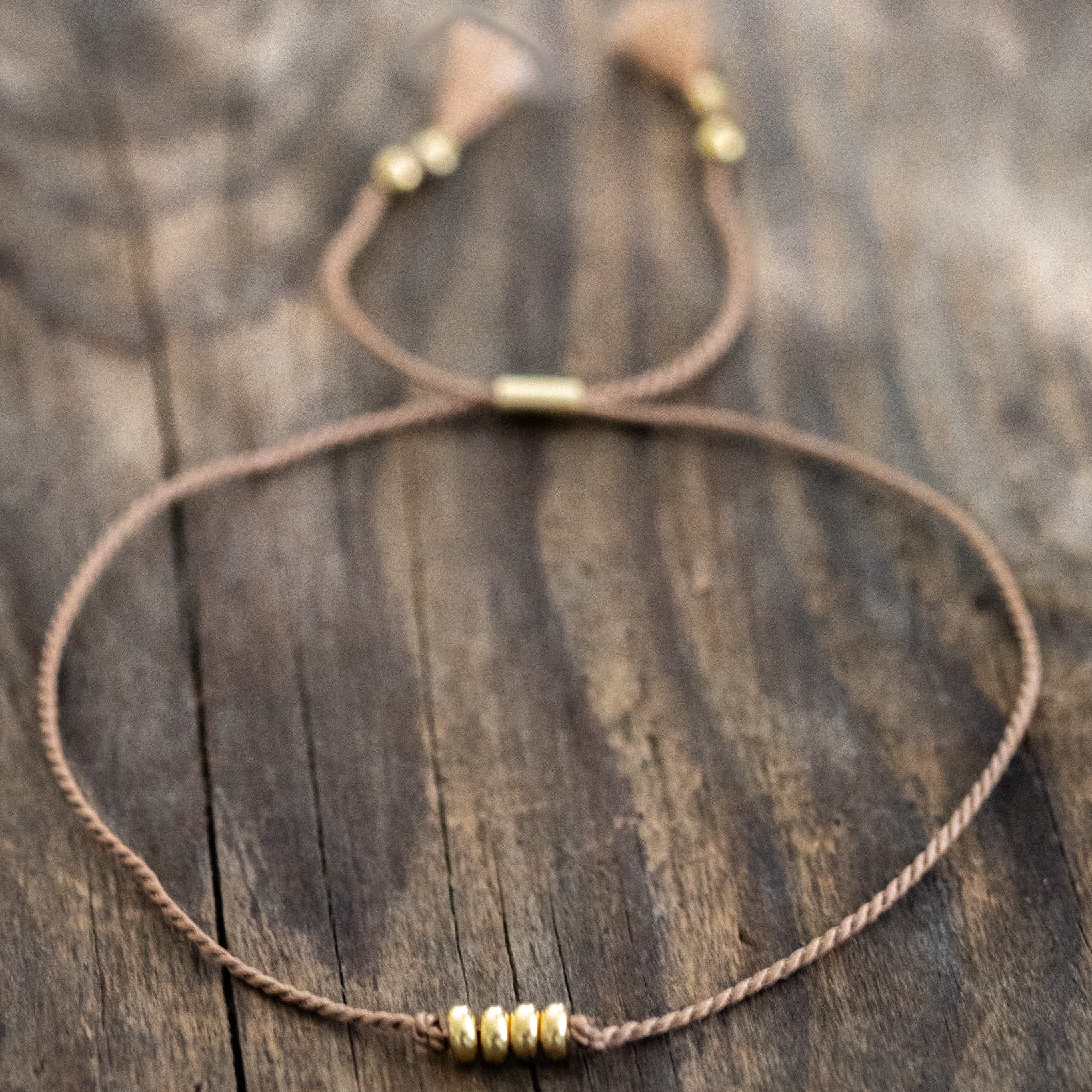 Ein dünnes, beiges Schnurarmband mit vier Goldperlen in der Mitte liegt auf braunem Holz.