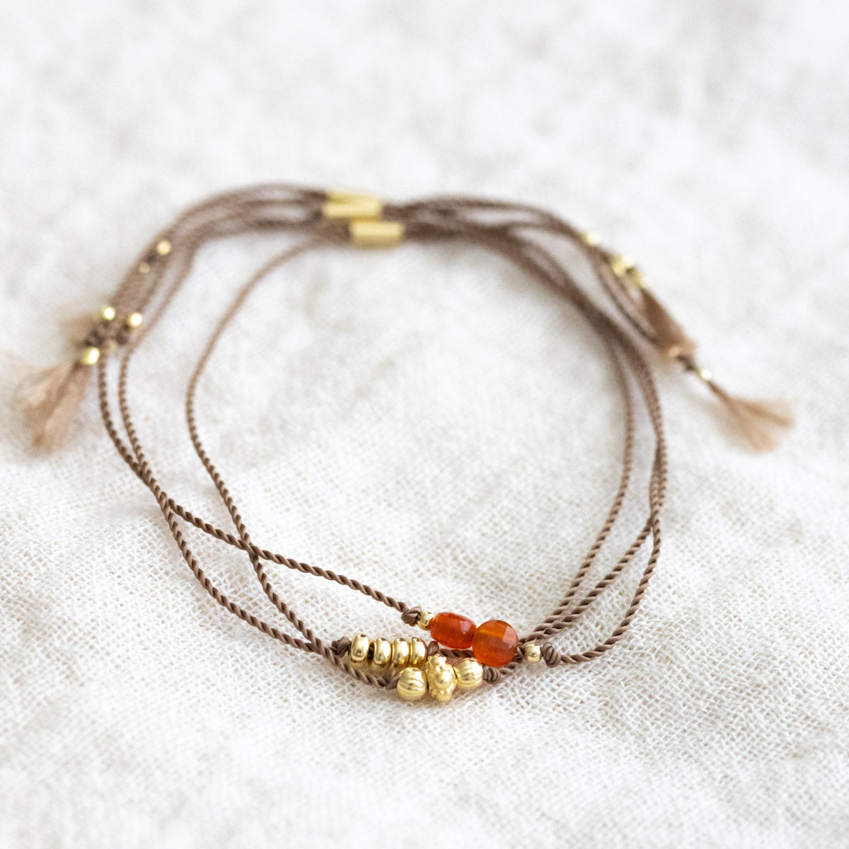 Drei unterschiedliche dünne, braune Schnurarmbänder, teilweise mit goldenen Perlen, teilweise mit orangem Karneol auf weißem Stoff ausgelegt.