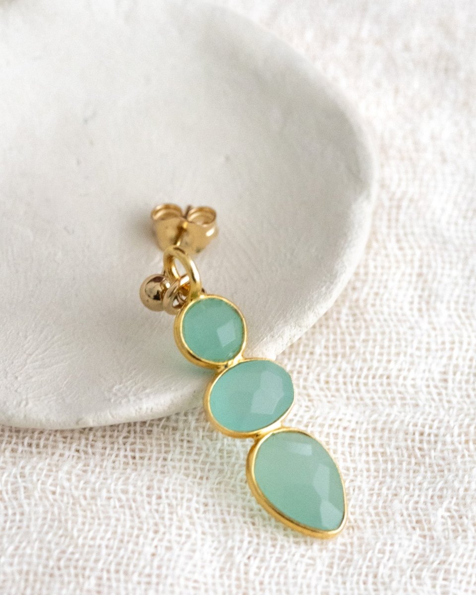 Ein langer, goldener Ohrring mit drei meeresfarbenen Aqua Chalcedonen lehnt an einem weißen Schälchen auf weißem Leinenstoff.