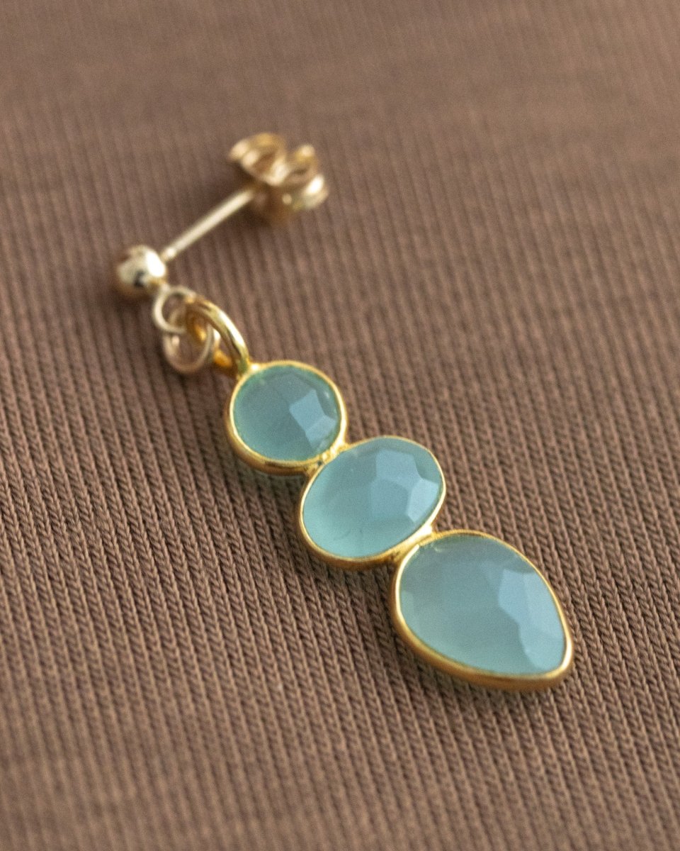 Ein langer, goldener Ohrring mit drei meeresfarbenen Aqua Chalcedonen liegt auf braunem Hintergrund.