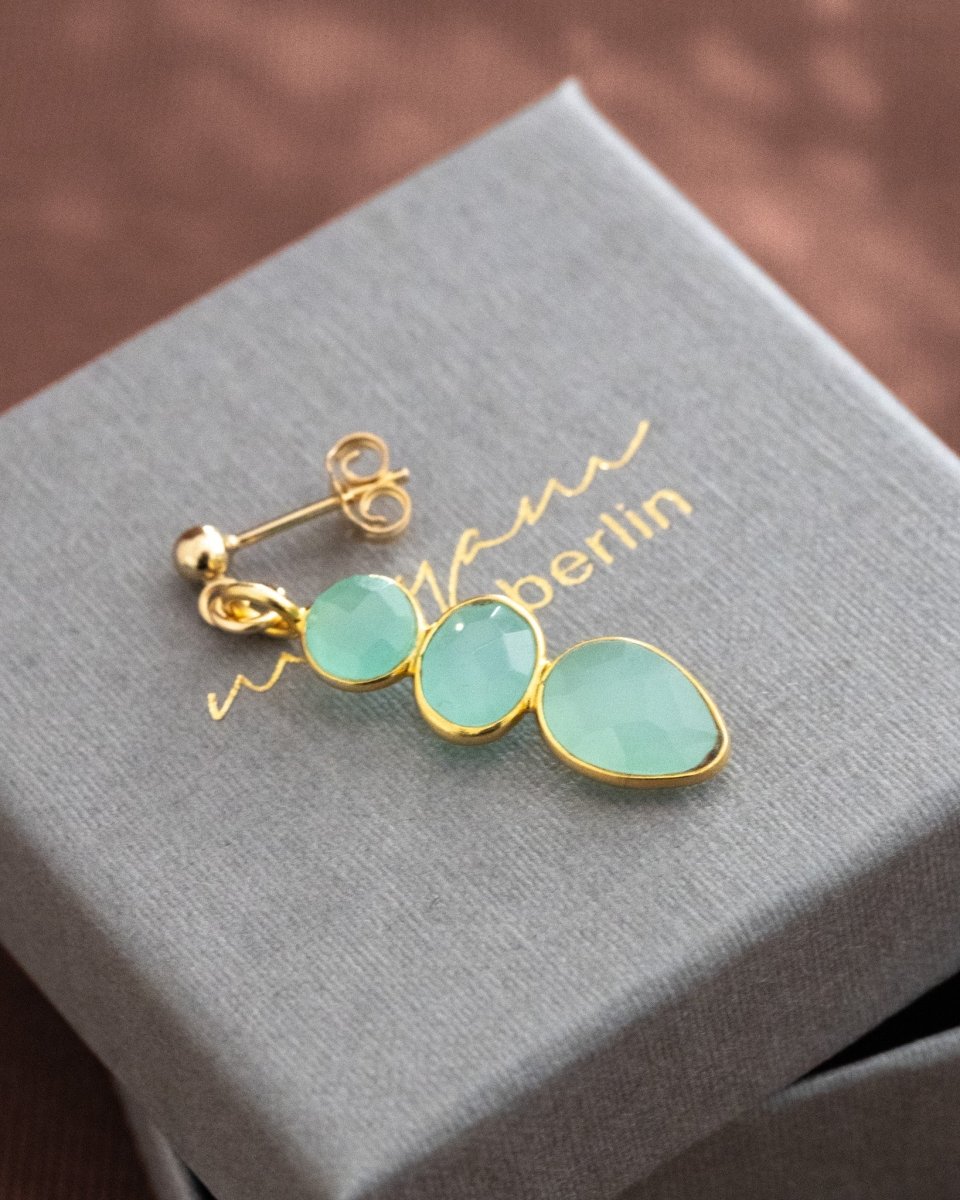 Ein langer, goldener Ohrring mit drei meeresfarbenen Aqua Chalcedonen liegt auf einem grauen Schmucketui.